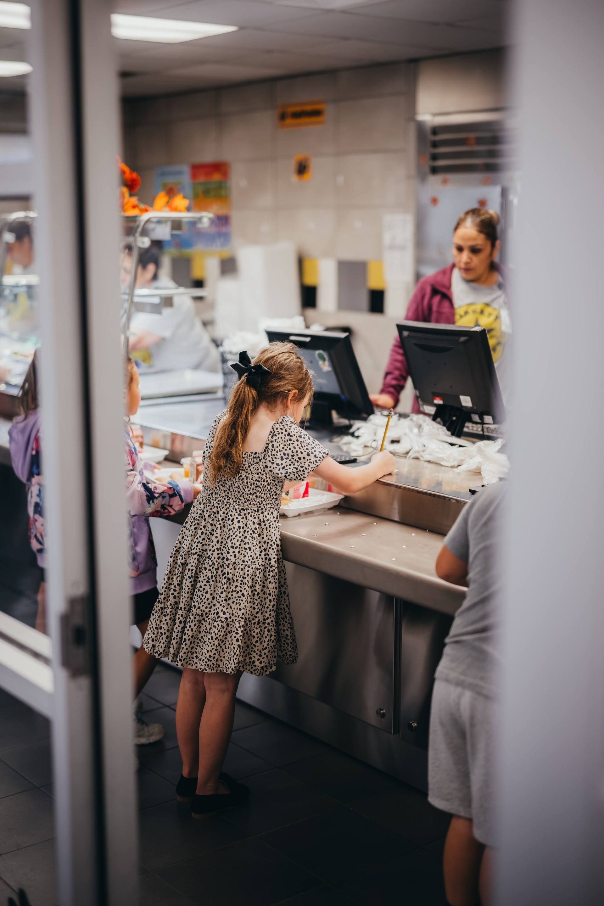 Children at a school cafeteria counter, girl reaching, worker standing behind the register.