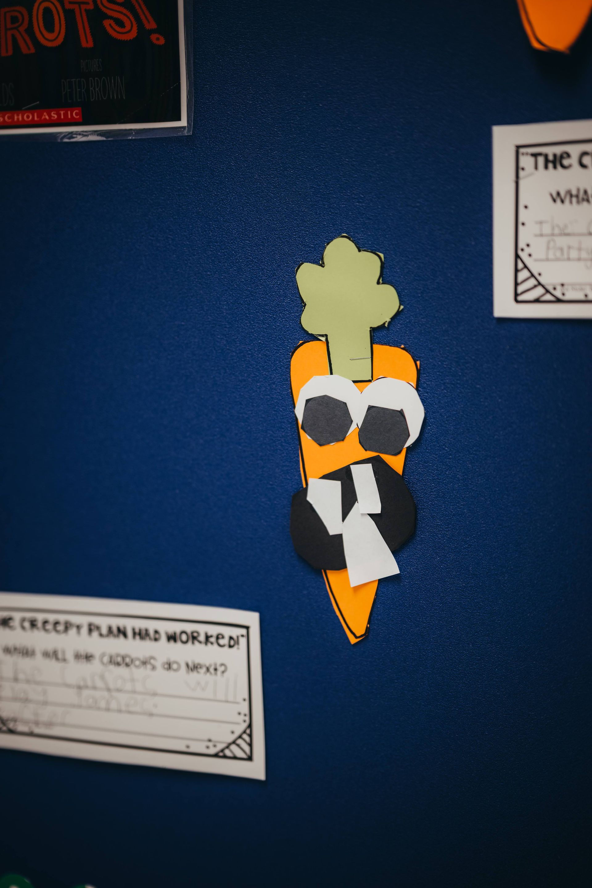 A cartoon carrot with googly eyes and a black tie is on a blue wall.