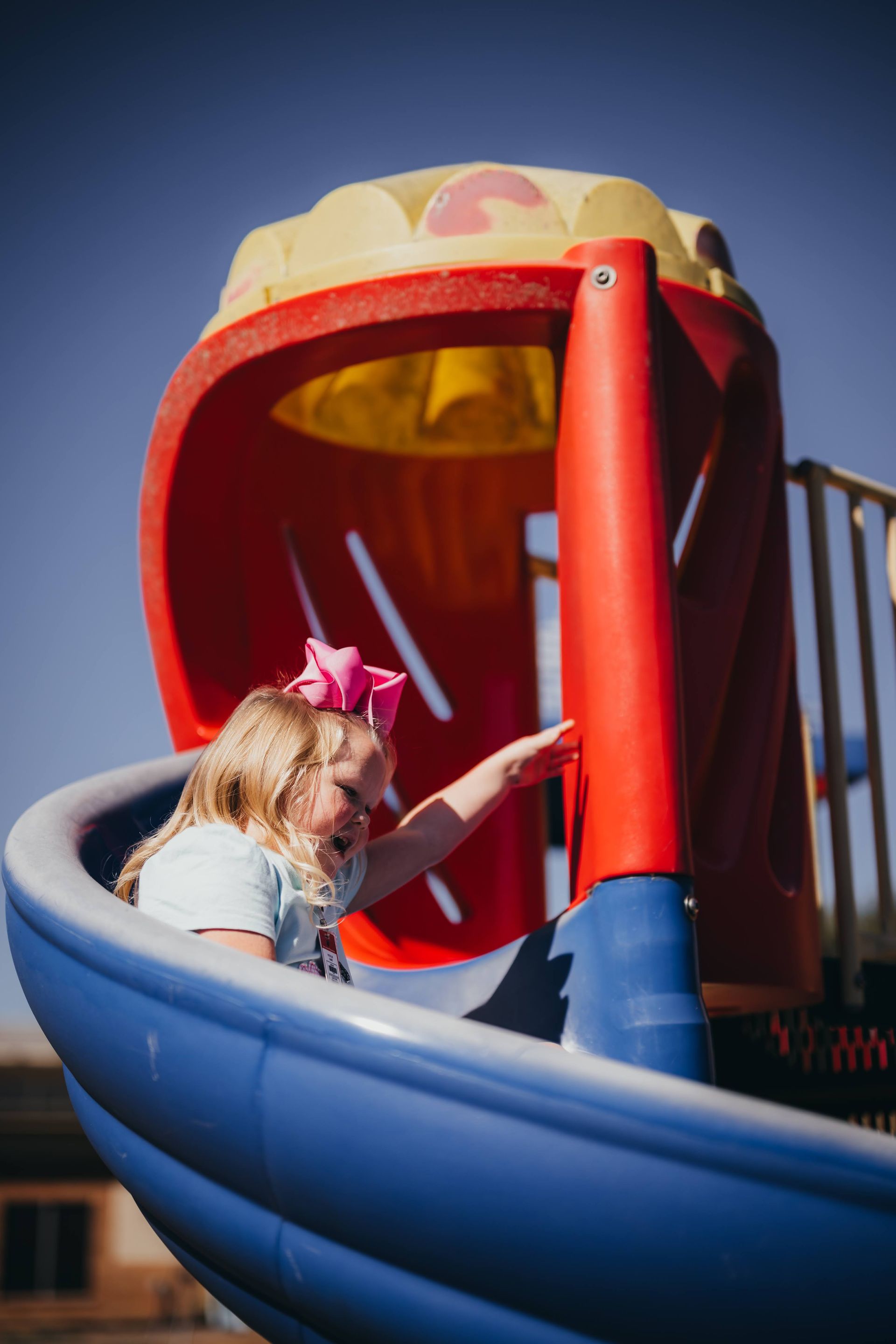Child on a blue and red playground slide, reaching out. Bright sky, pink bow.