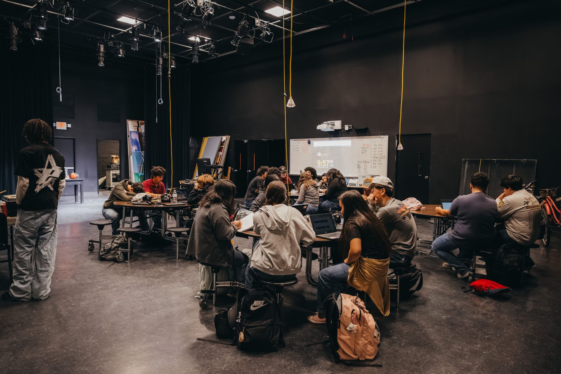A group of people sitting around a table in a dark room with a projector screen, working on computers.