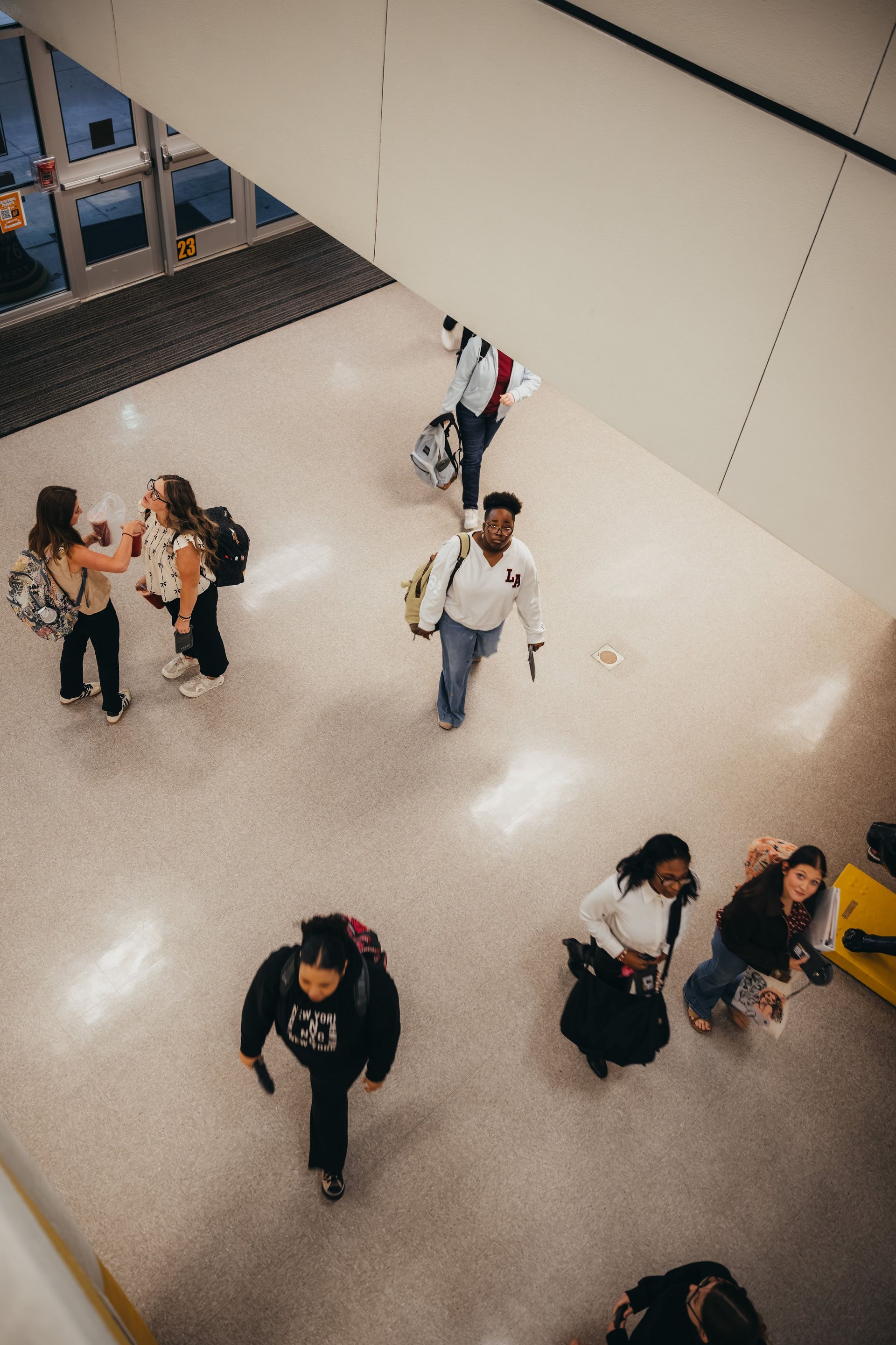 People walking in a brightly lit airport terminal, some with luggage, near a set of doors.