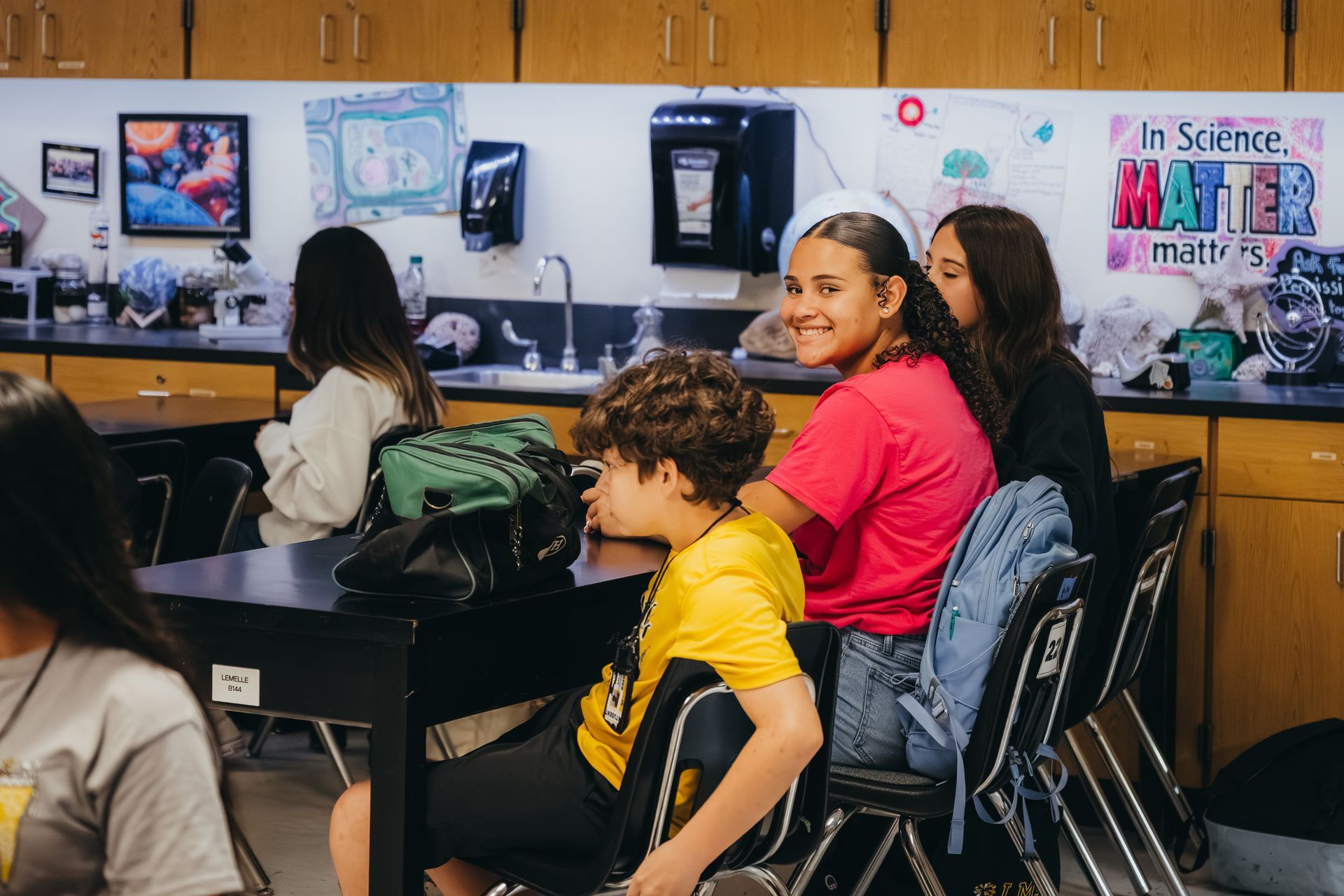Students in a classroom. One smiles at the camera, wearing pink, others in yellow and black. Desks and lab equipment.