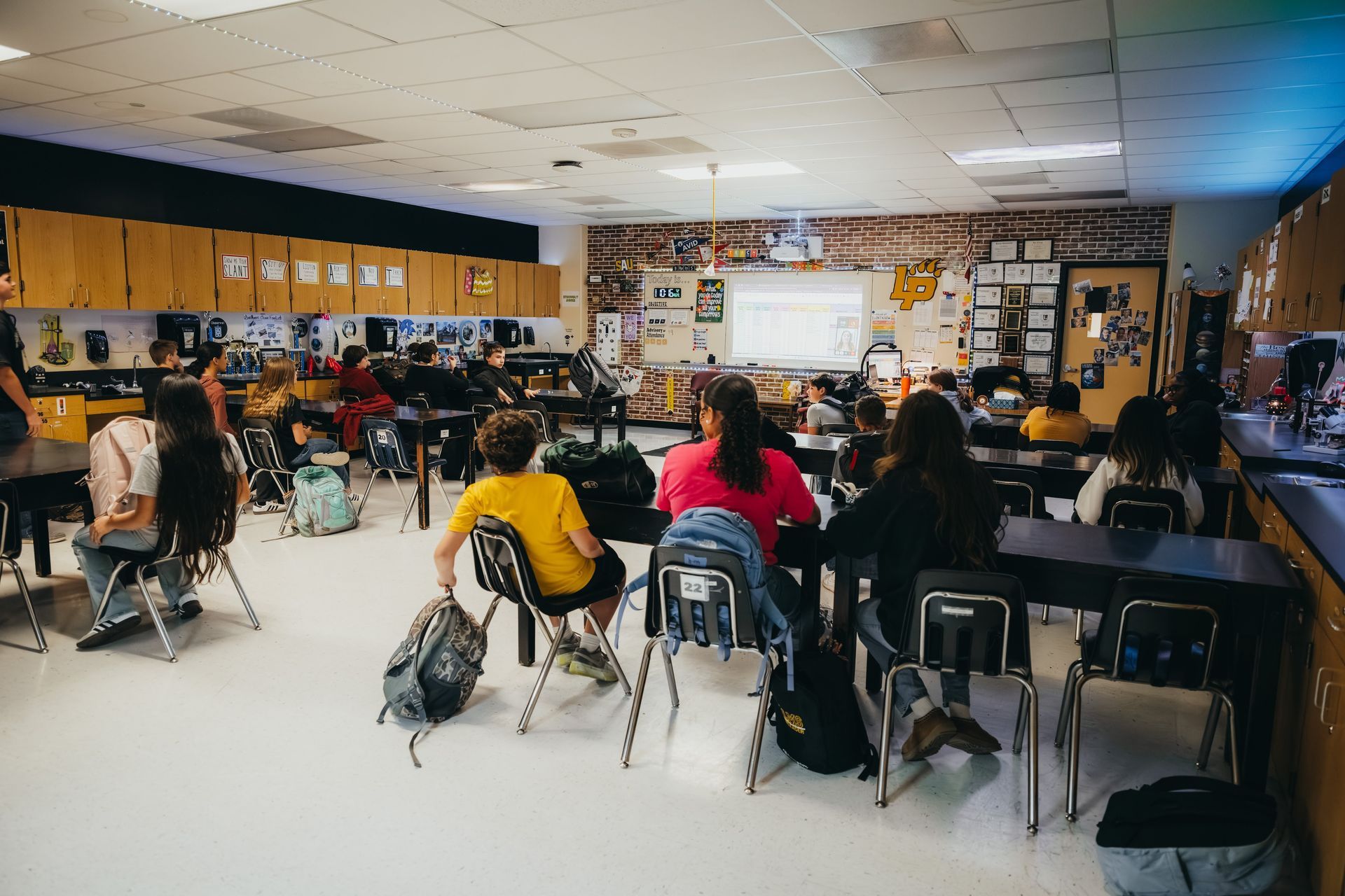 Classroom with students seated at desks, facing forward. Display screen on the wall.