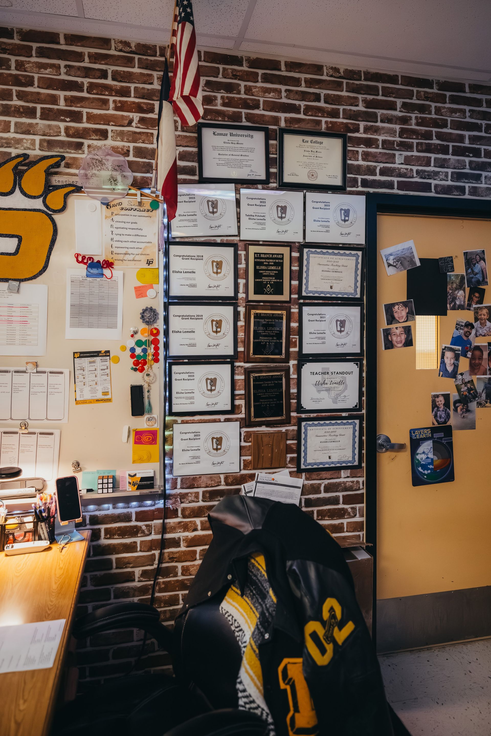 A classroom corner with brick wall, awards, American flag, desk, and a door with photos.