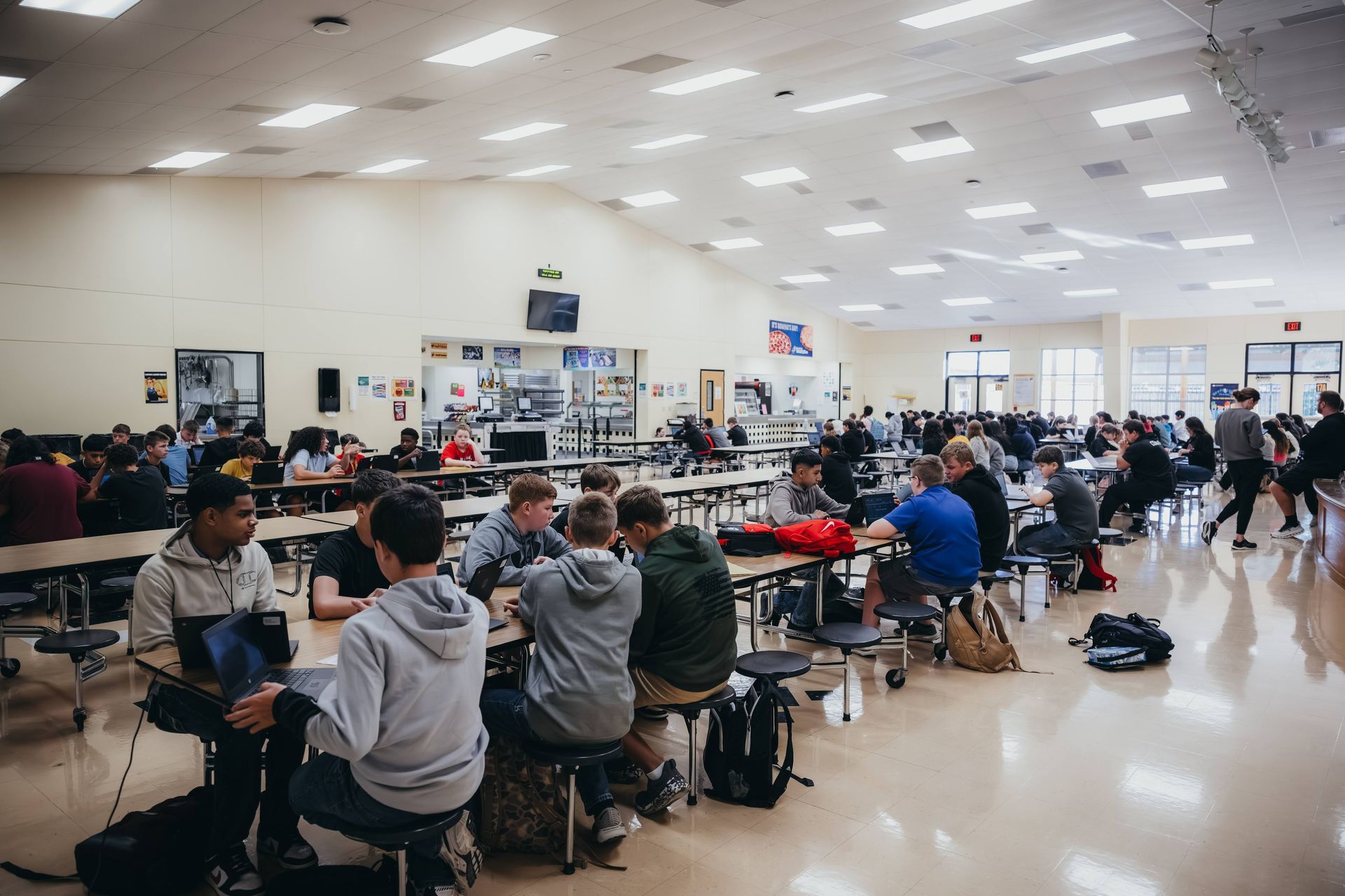 School cafeteria with students seated at tables, some talking and eating. Bright, open space.