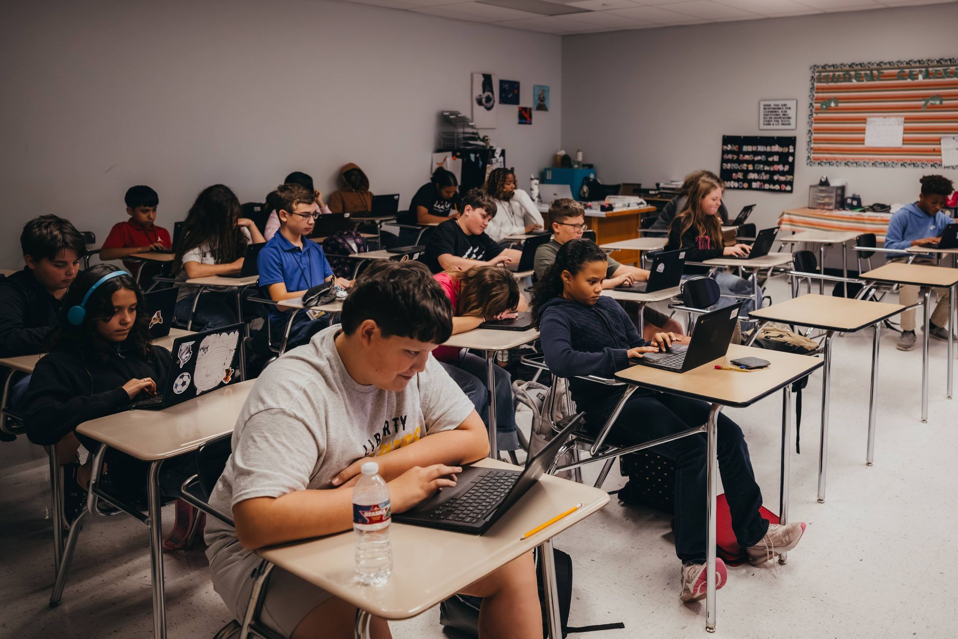 Students seated at desks in a classroom, using laptops. One student has a water bottle.