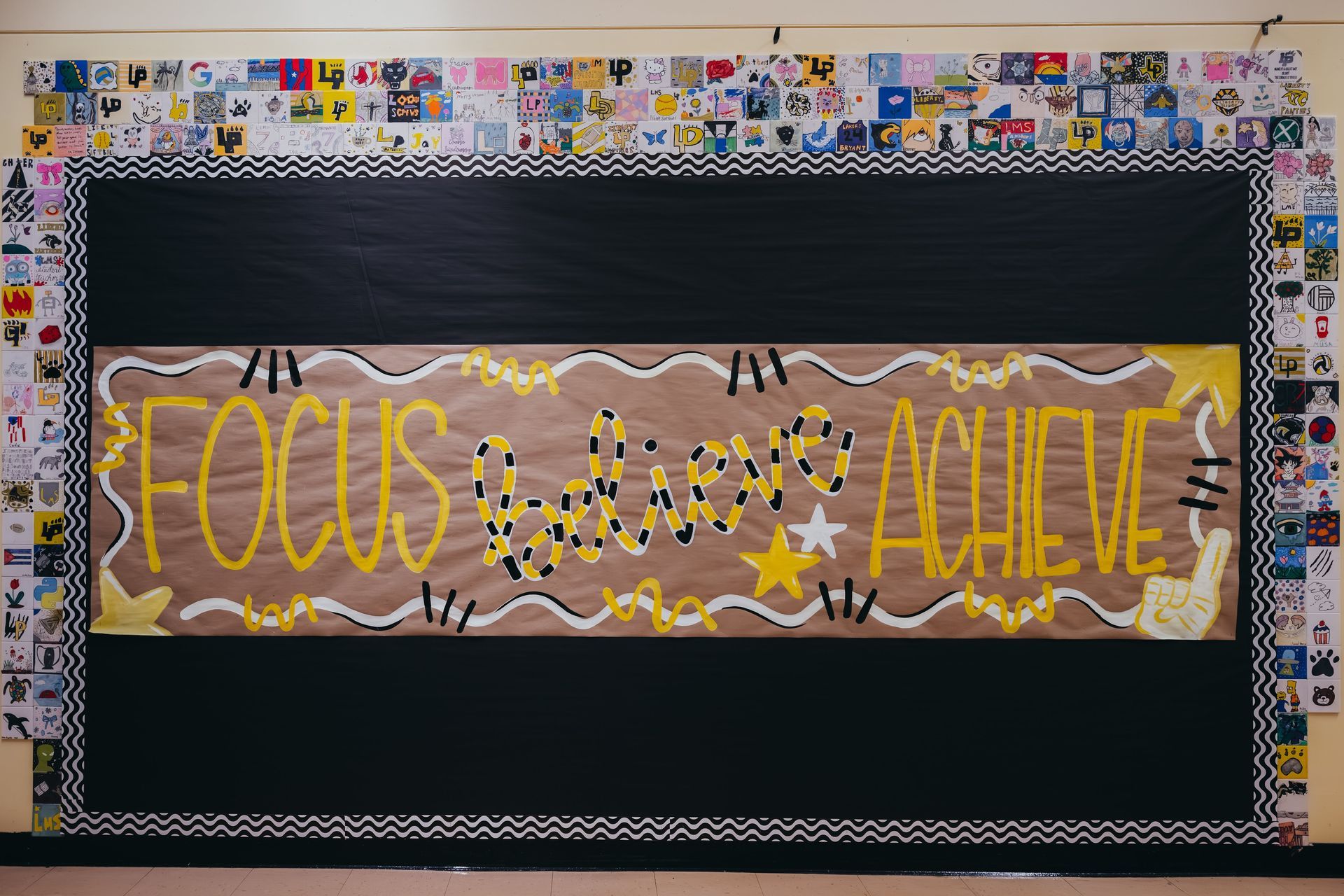 Bulletin board with FOCUS BELIEVE ACHIEVE in yellow on a brown paper, framed in black and surrounded by student artwork.