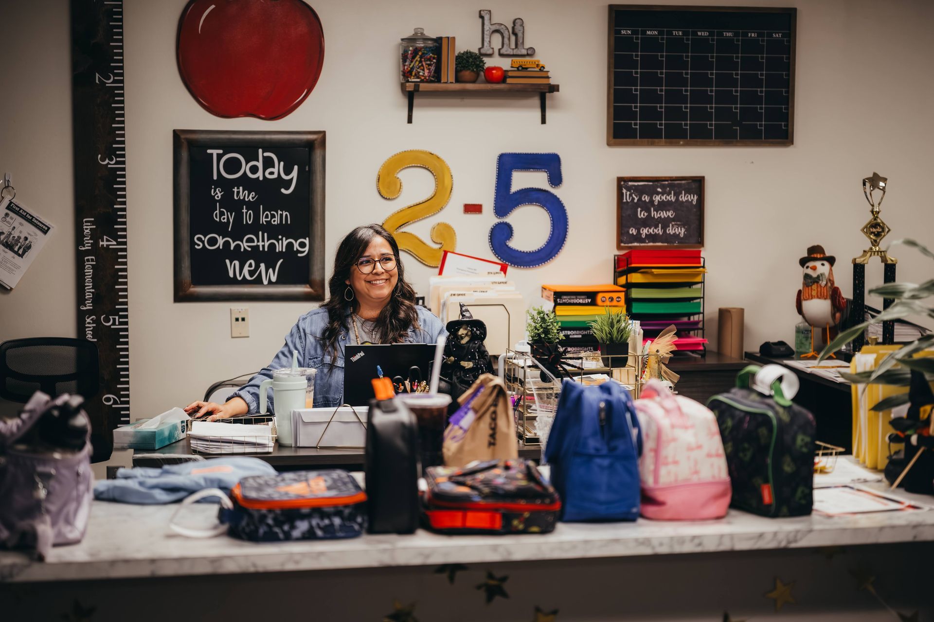 Teacher at desk in a colorful classroom, surrounded by school supplies and decorations.