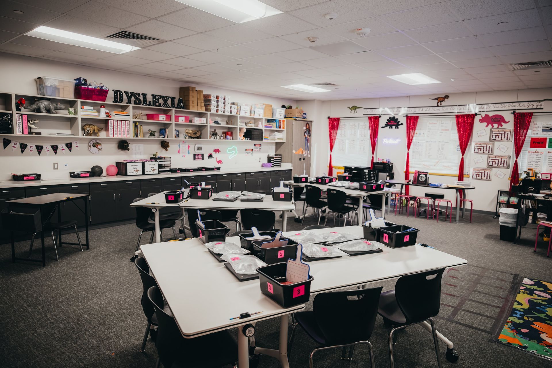 Classroom interior, desks with laptops, storage shelves, red curtains, organized.