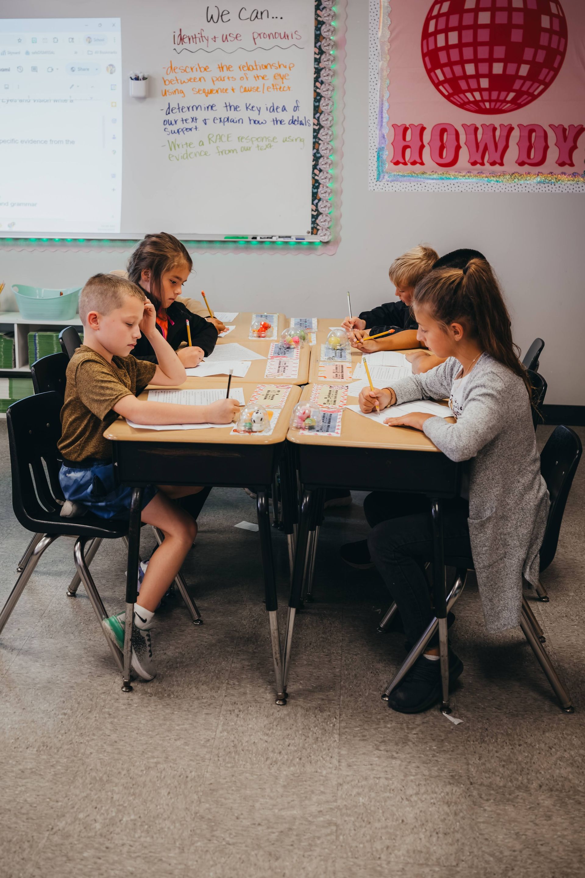 Four children seated at desks in a classroom, writing. A disco ball sign hangs on a wall.