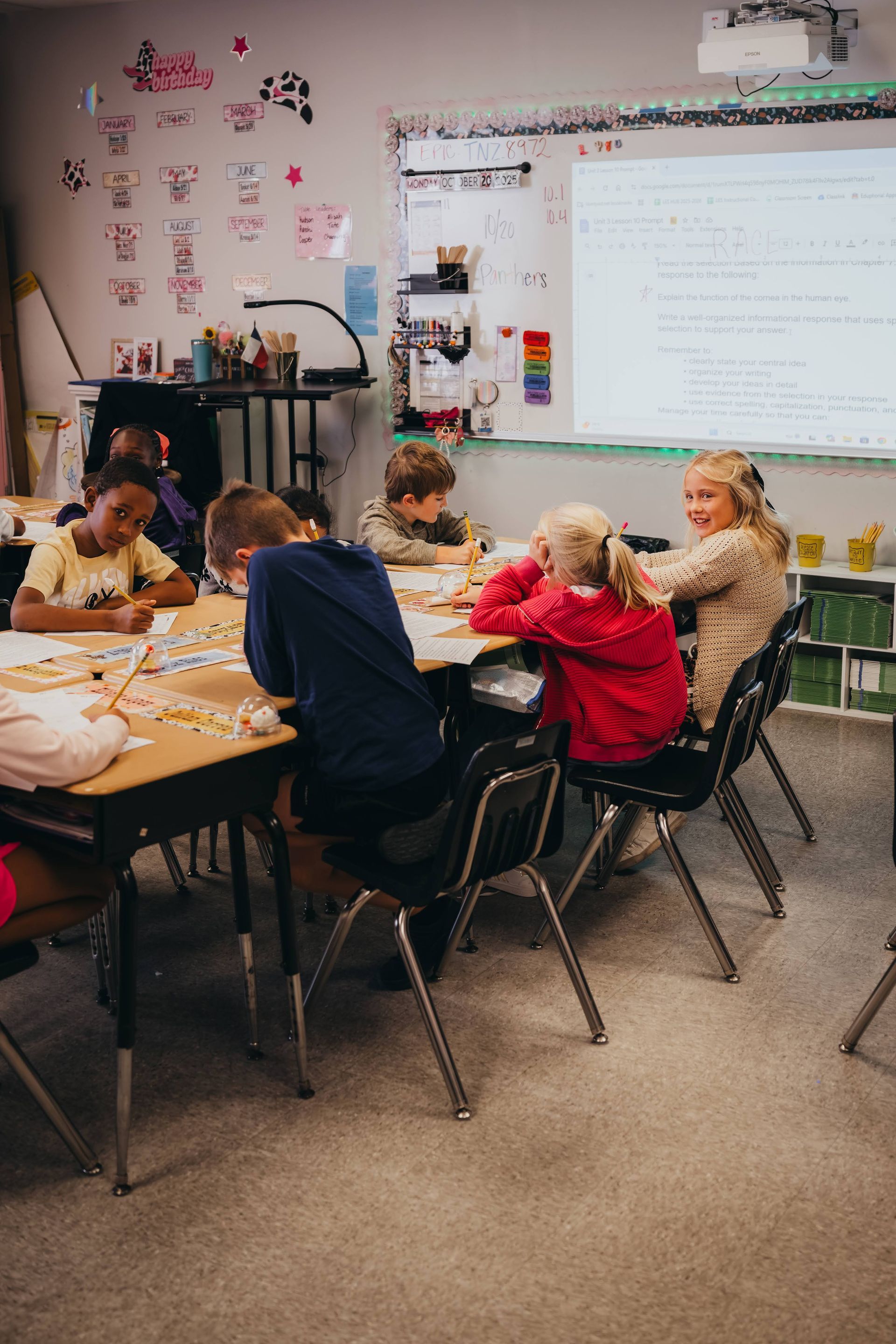 Children seated around a table in a classroom work on a project. A whiteboard and decorations are in the background.