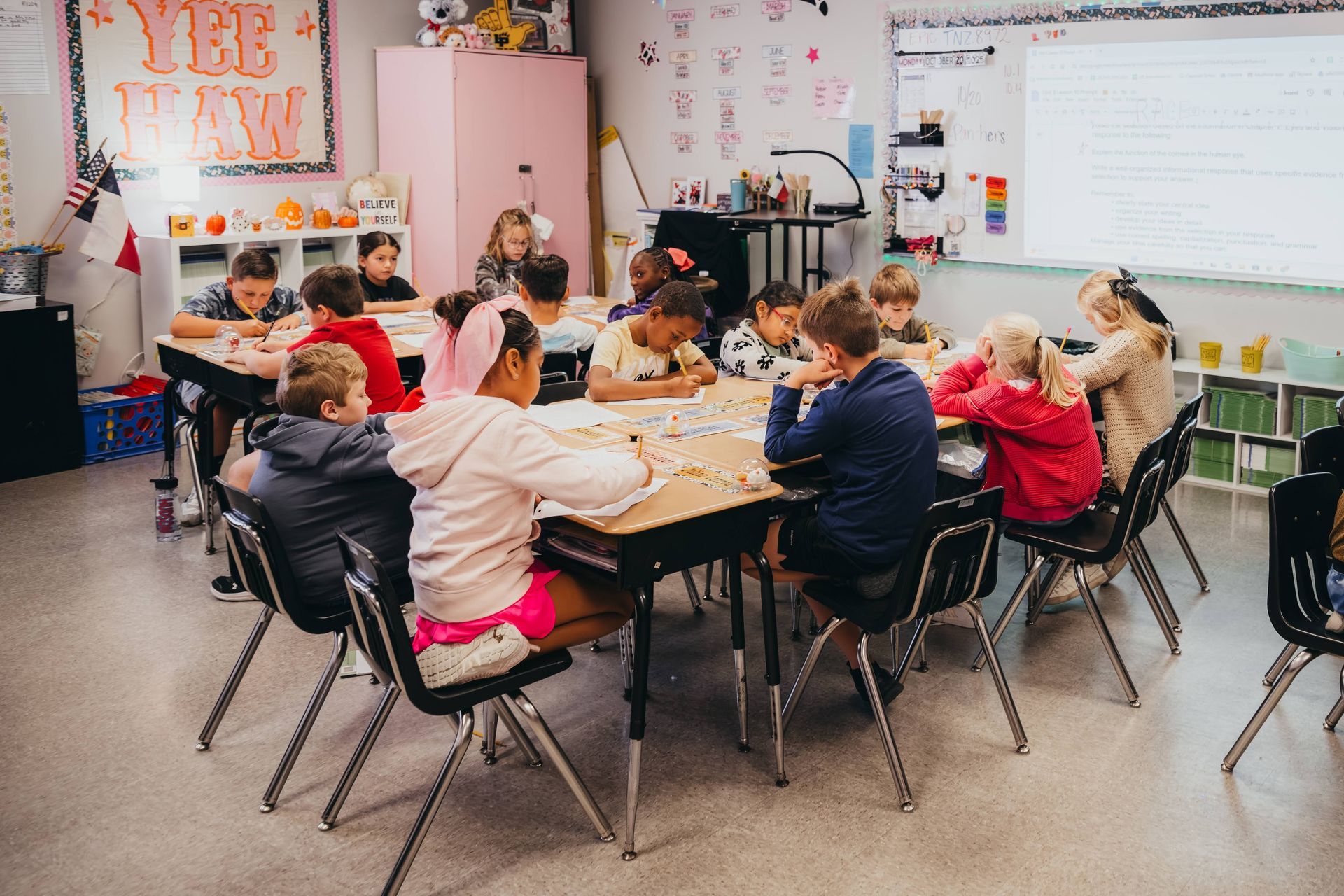 Children work at a table in a classroom, focused on an activity. The setting is bright with colorful decorations.