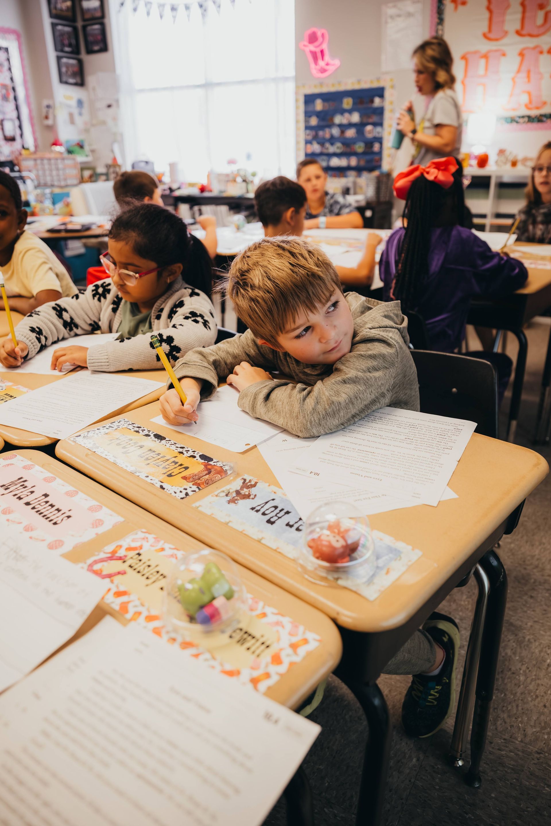 Children working at desks in a classroom, one boy looking to the side, wearing a knit hat.