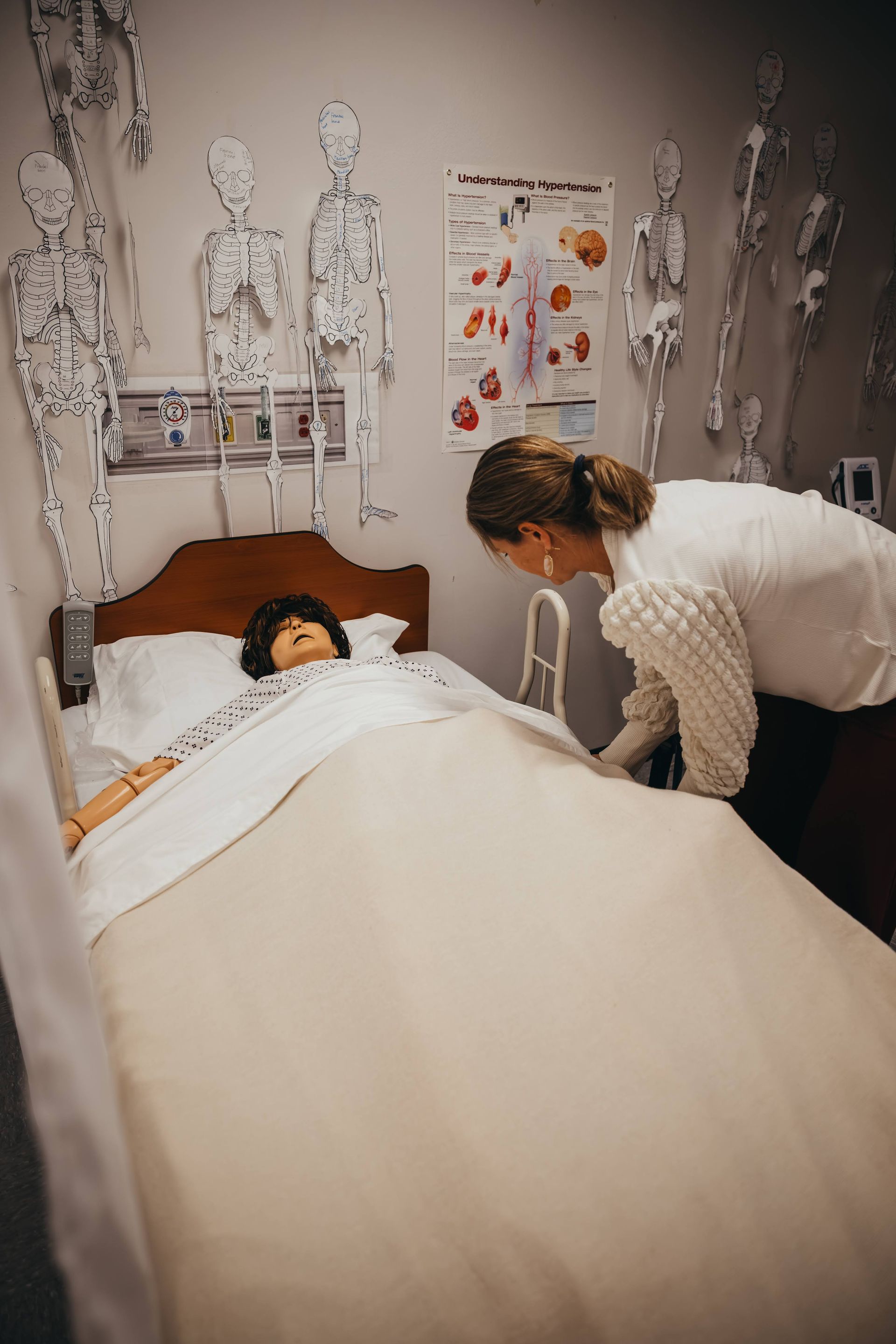 Woman in scrubs tending to a bed-ridden medical mannequin in a hospital room, skeleton diagrams on wall.