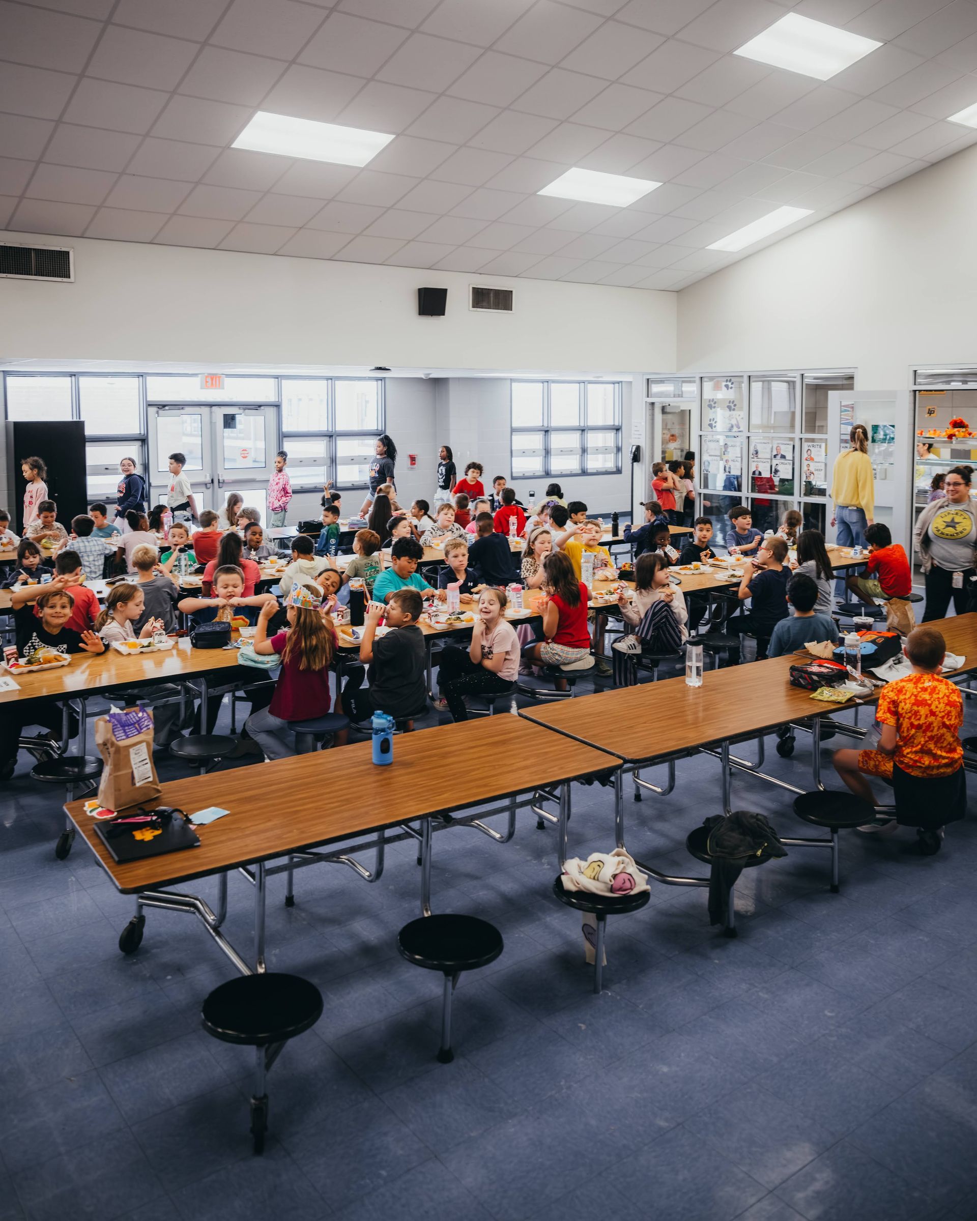 School cafeteria scene with students eating lunch at tables. Bright, open space.