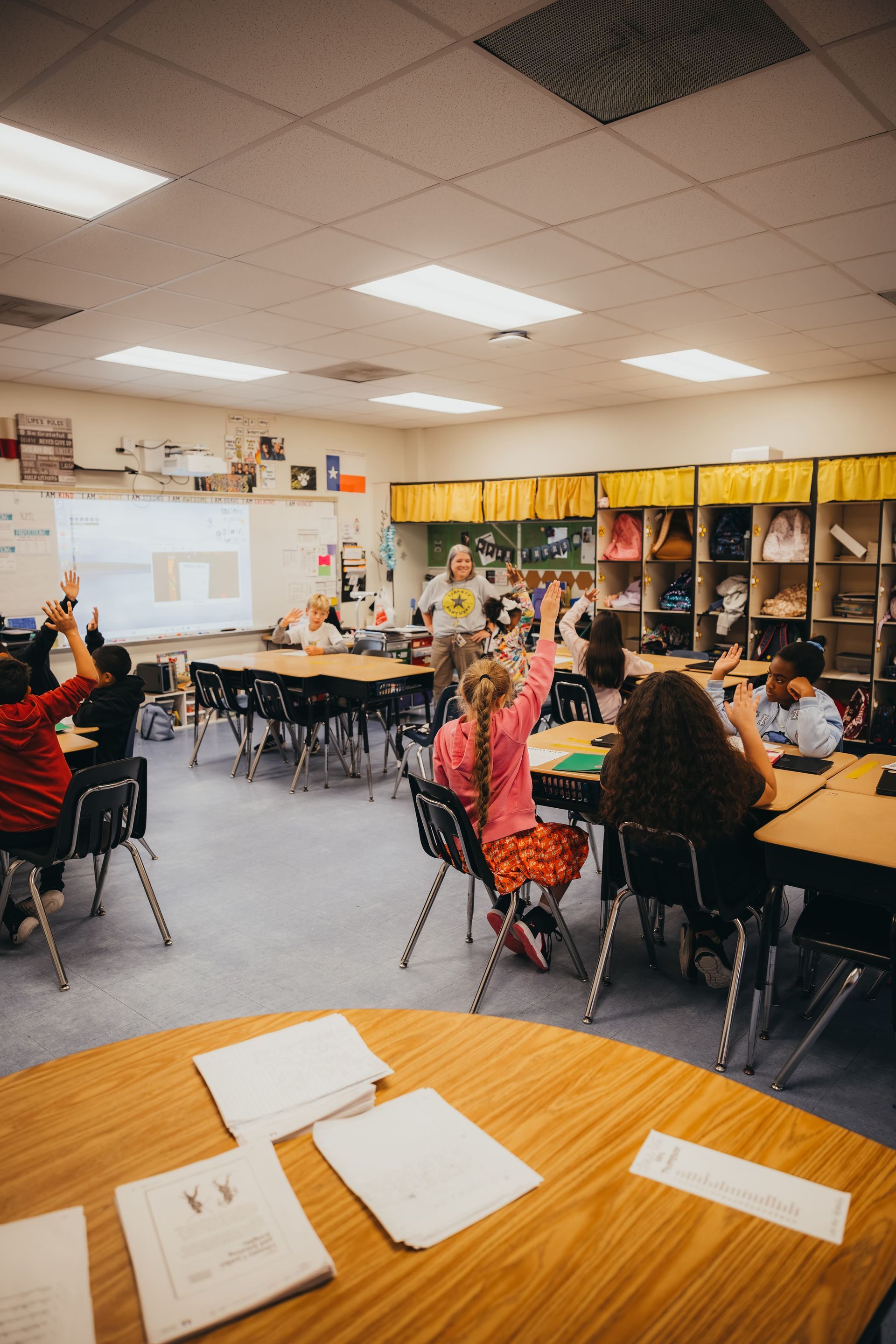 Students raising hands in a classroom, teacher at the whiteboard, desks arranged around the room.