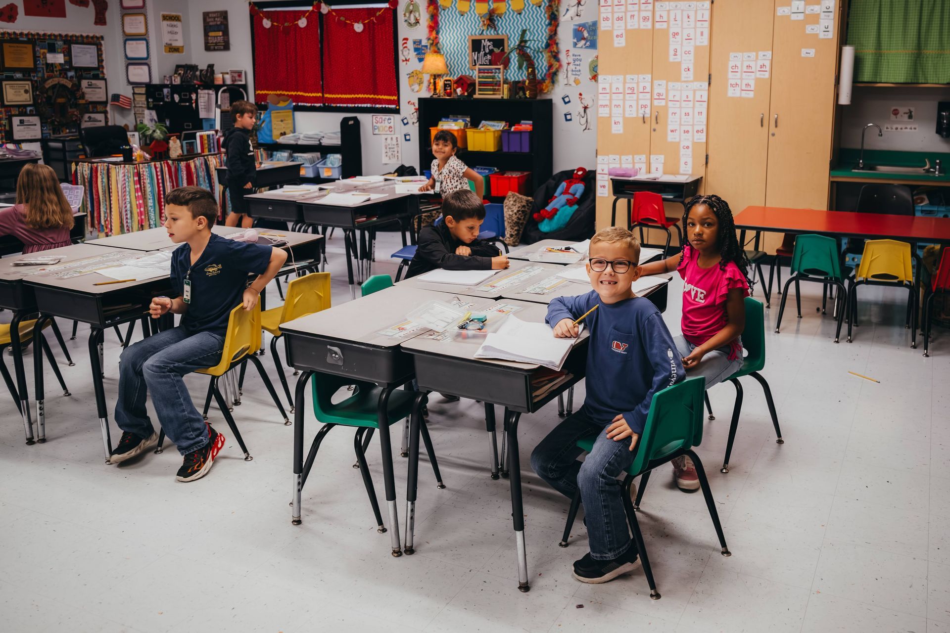 Children in a classroom, sitting at desks. Some are looking at the camera and smiling.