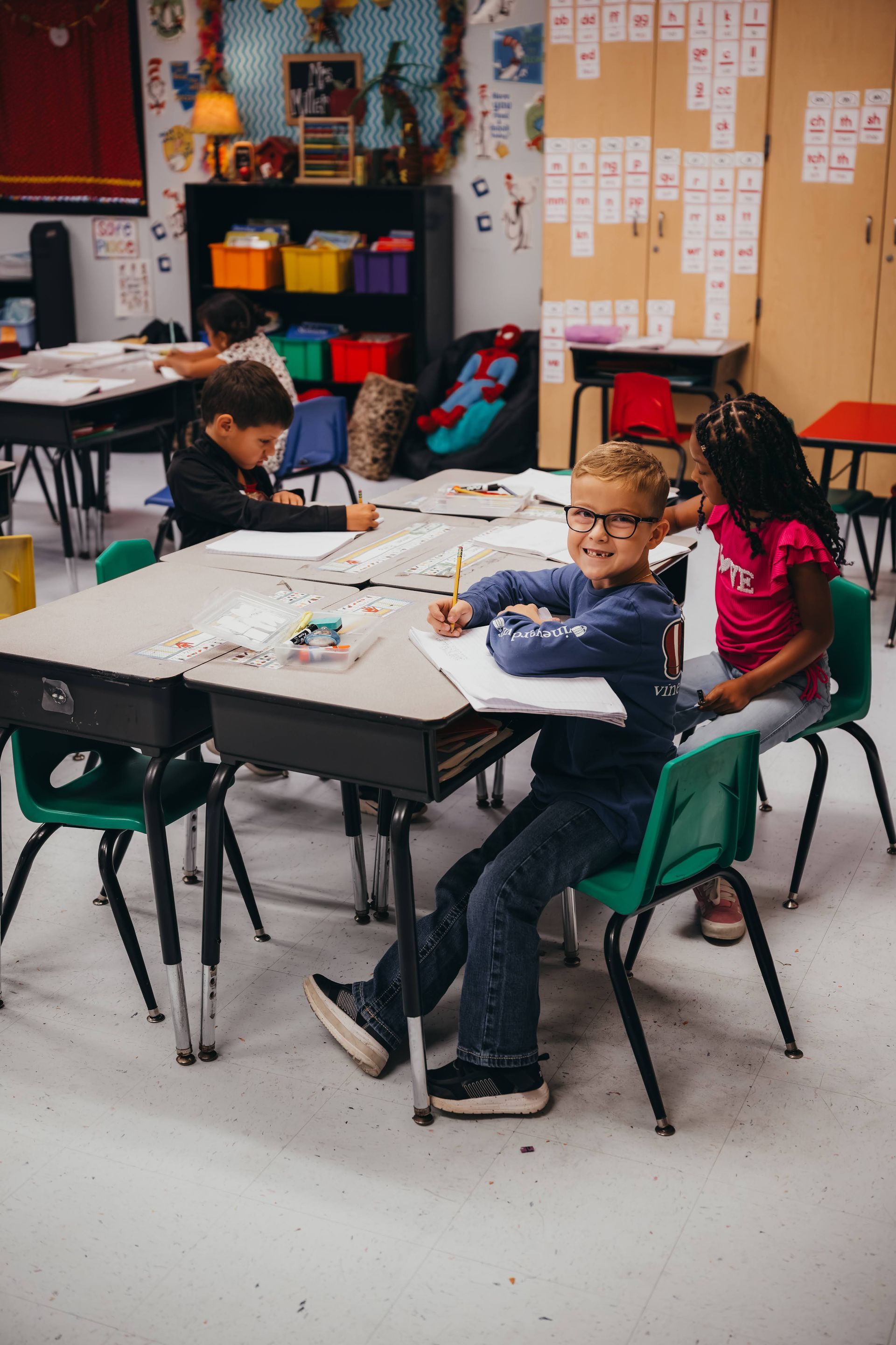 Three students at a desk in a classroom, one smiling. Colorful shelves and whiteboards in the background.