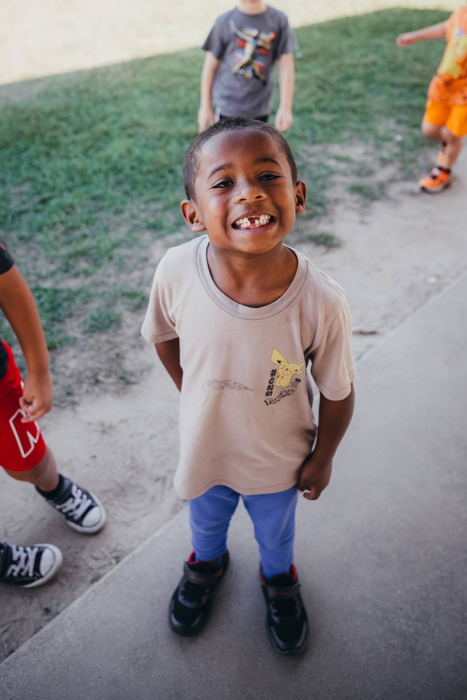 Smiling child in a beige shirt and blue pants, with other children playing in the background.