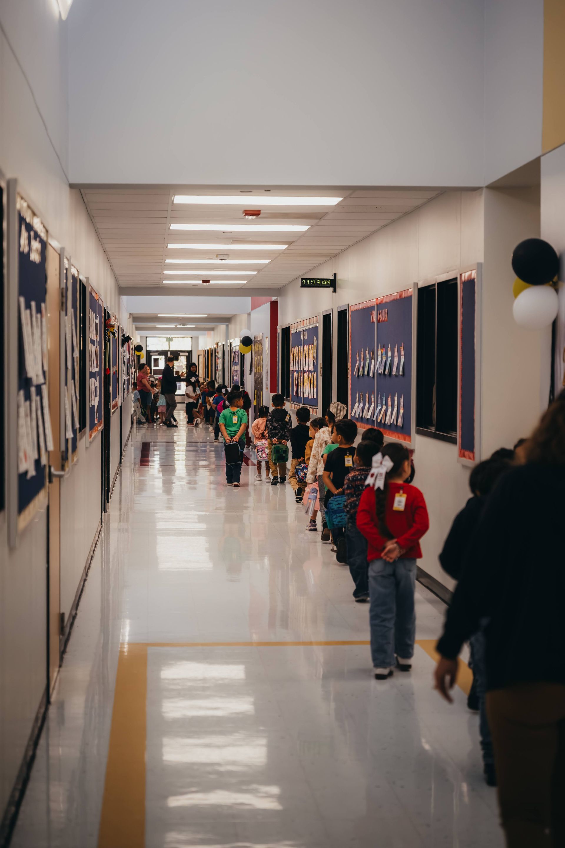 Children line a school hallway, waiting. Display boards line the walls, balloons at the end.