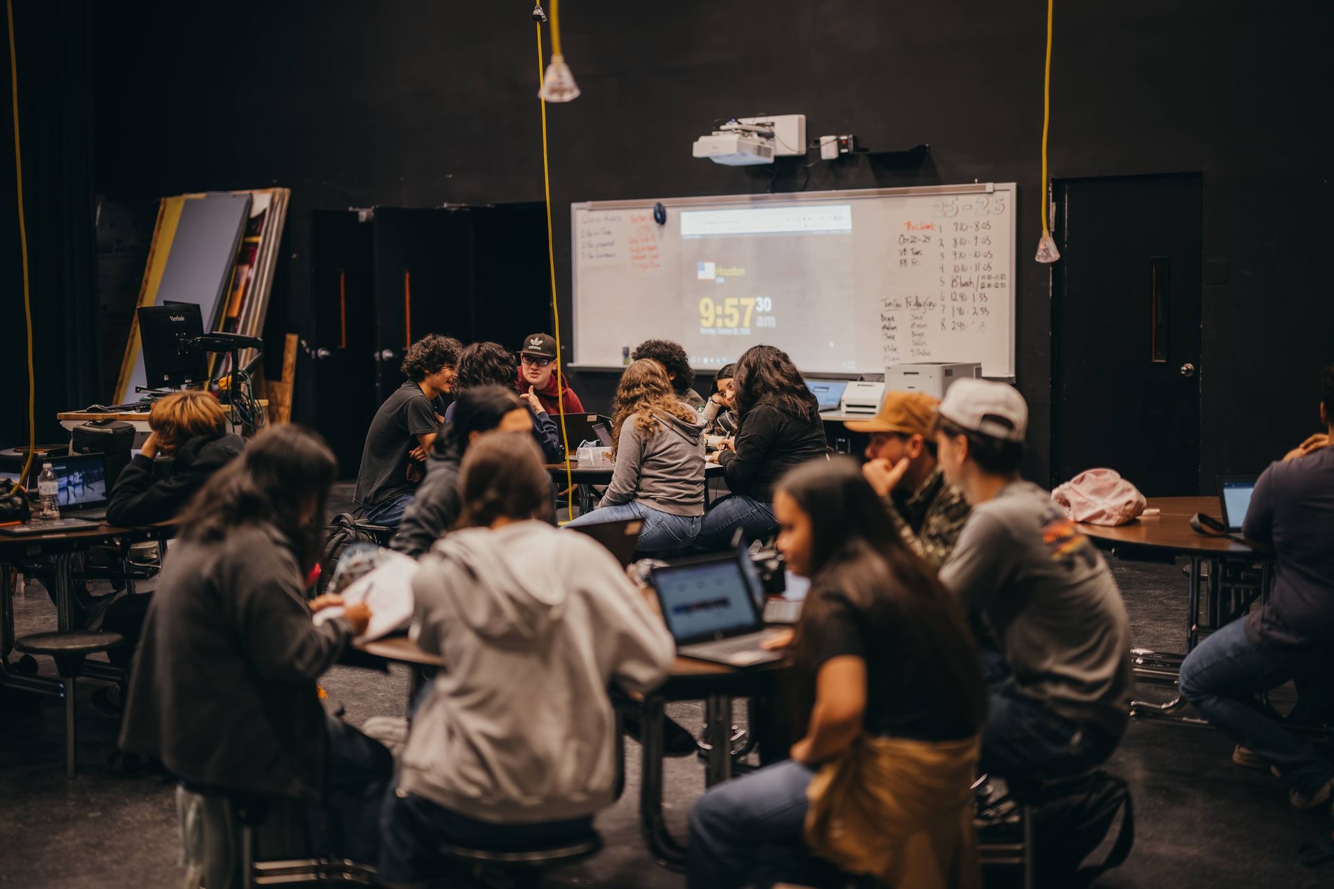 Students clustered around tables in a dimly lit room, some with laptops, whiteboard in background.