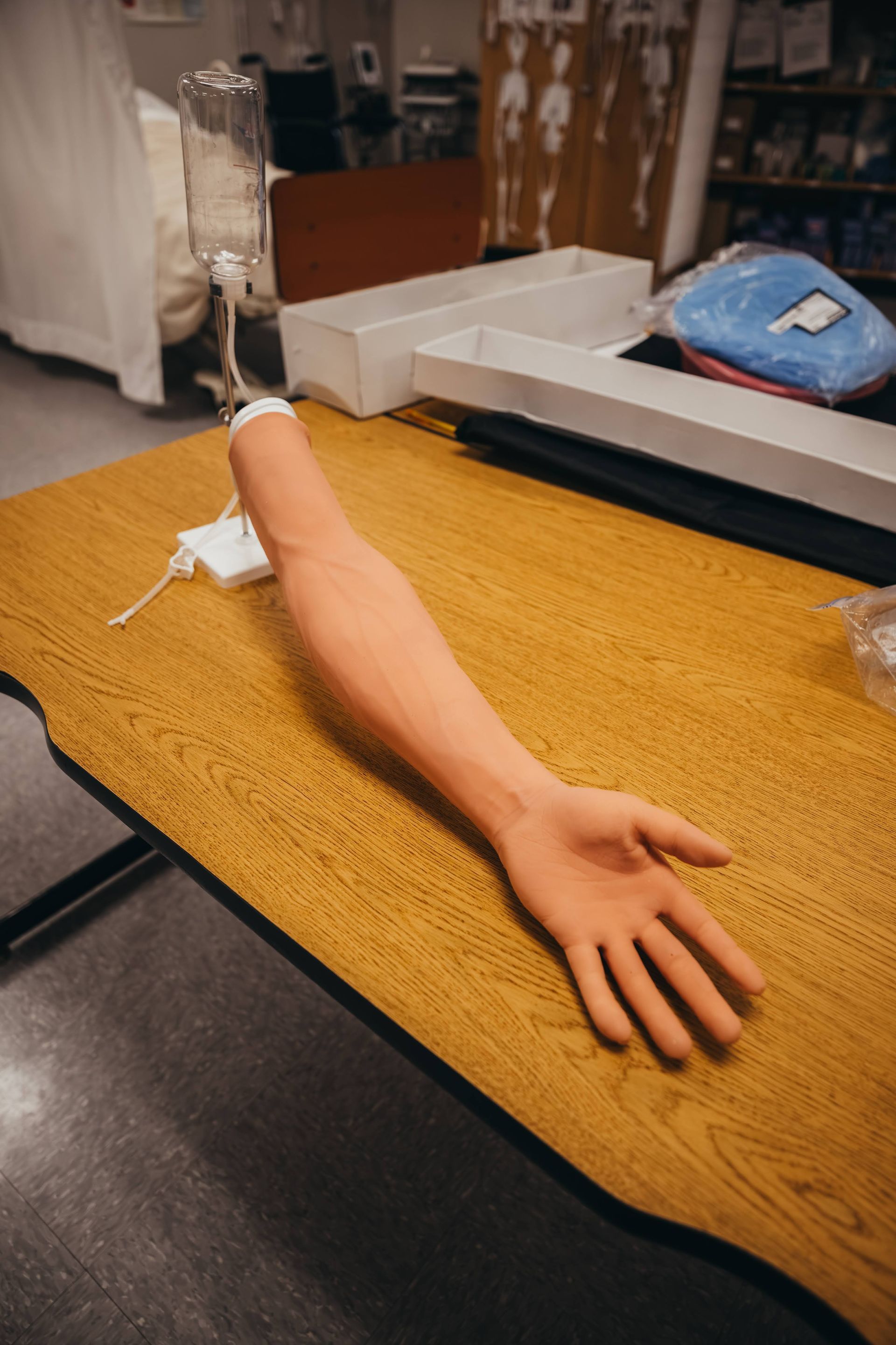 Medical arm model on a table, connected to IV bag. Training room setting, brown wooden table.