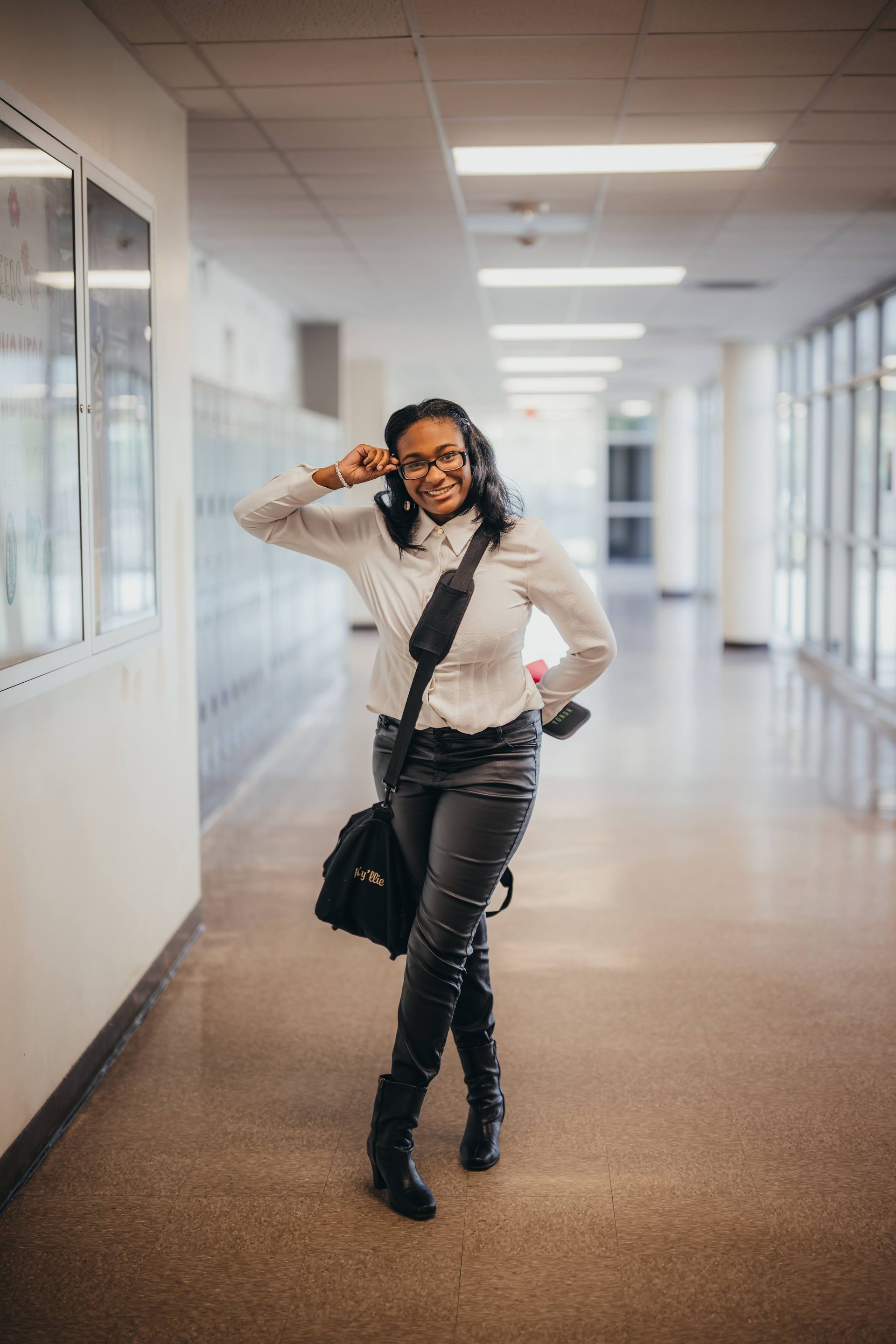 Person in a white shirt and black pants adjusts glasses, walking down a school hallway with a bag.