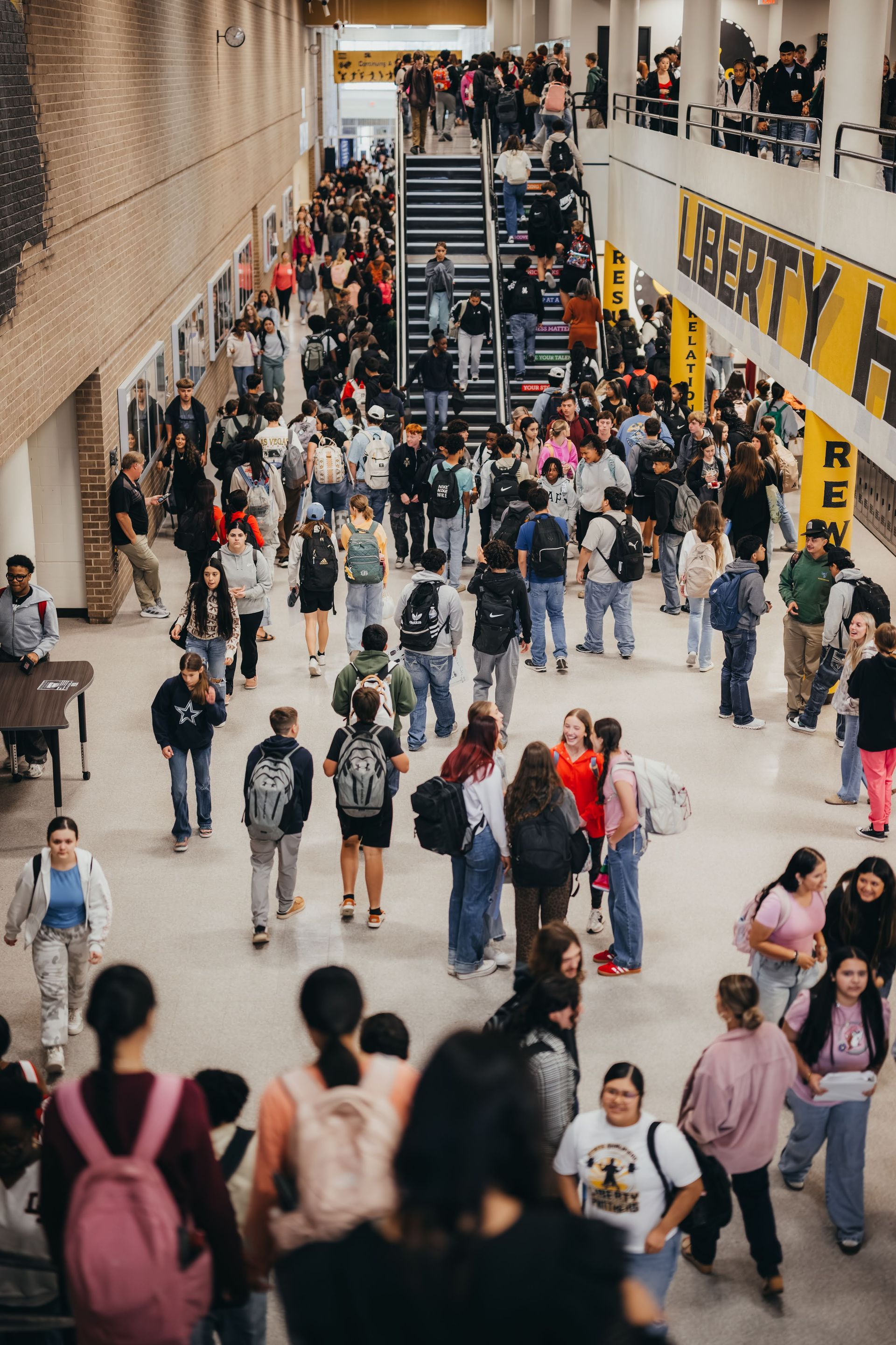 Students in a crowded high school hallway, walking and talking near stairs.