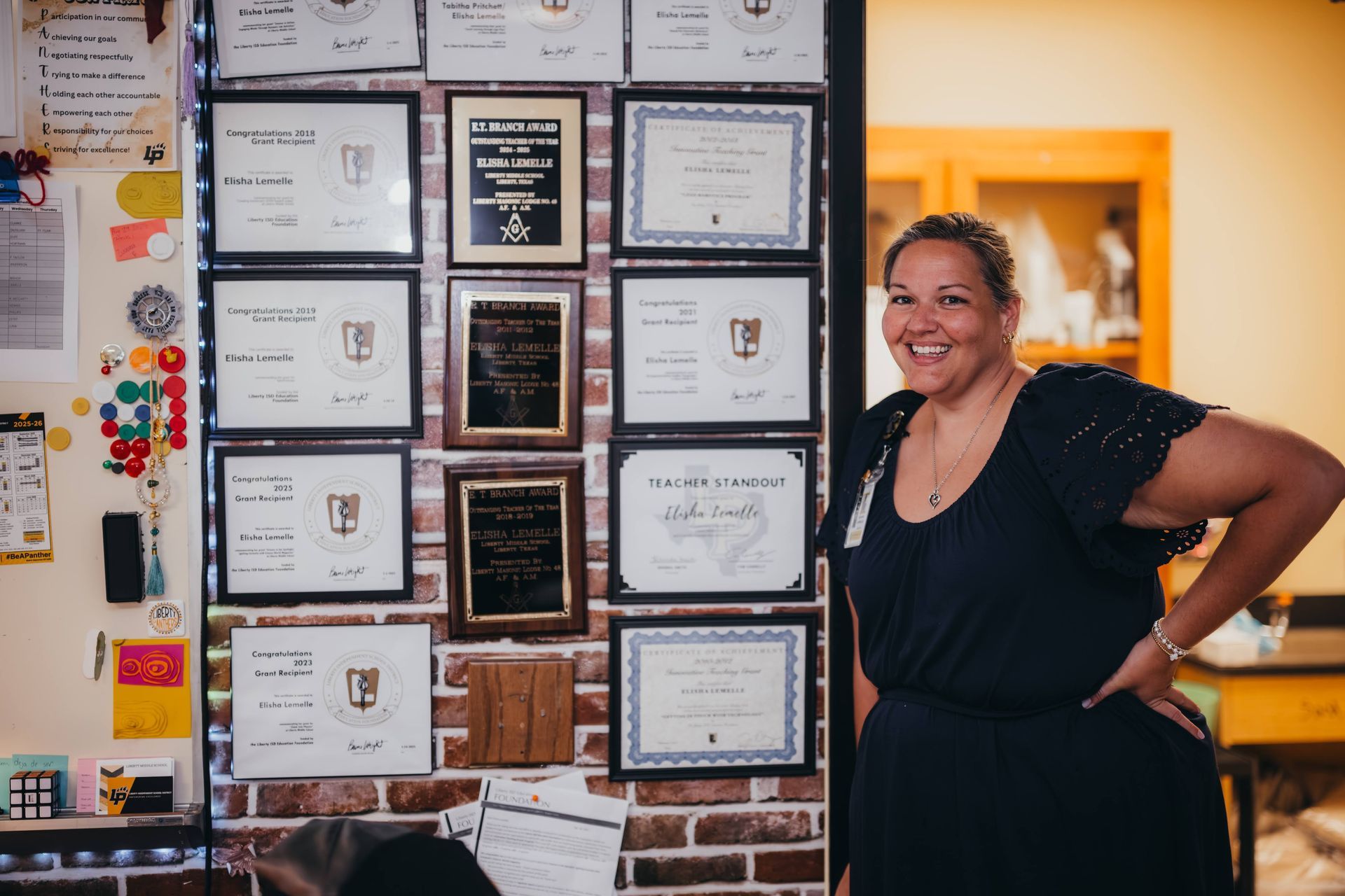 Woman smiling next to a wall covered in framed certificates. She wears a dark dress, hand on hip. Brick wall.
