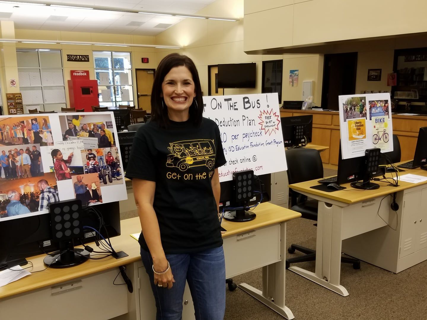 Woman in black shirt stands by posters in a library, smiling. Desks with computers behind.