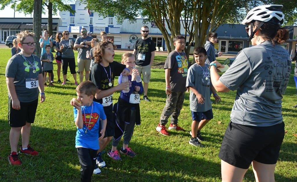 Children in running gear at a race, being instructed by a person wearing a helmet, outdoors, daytime.