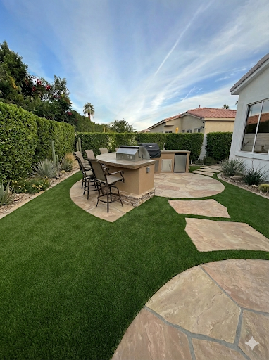 Outdoor kitchen area with built-in grill, bar, and seating on stone patio; green lawn and blue sky.