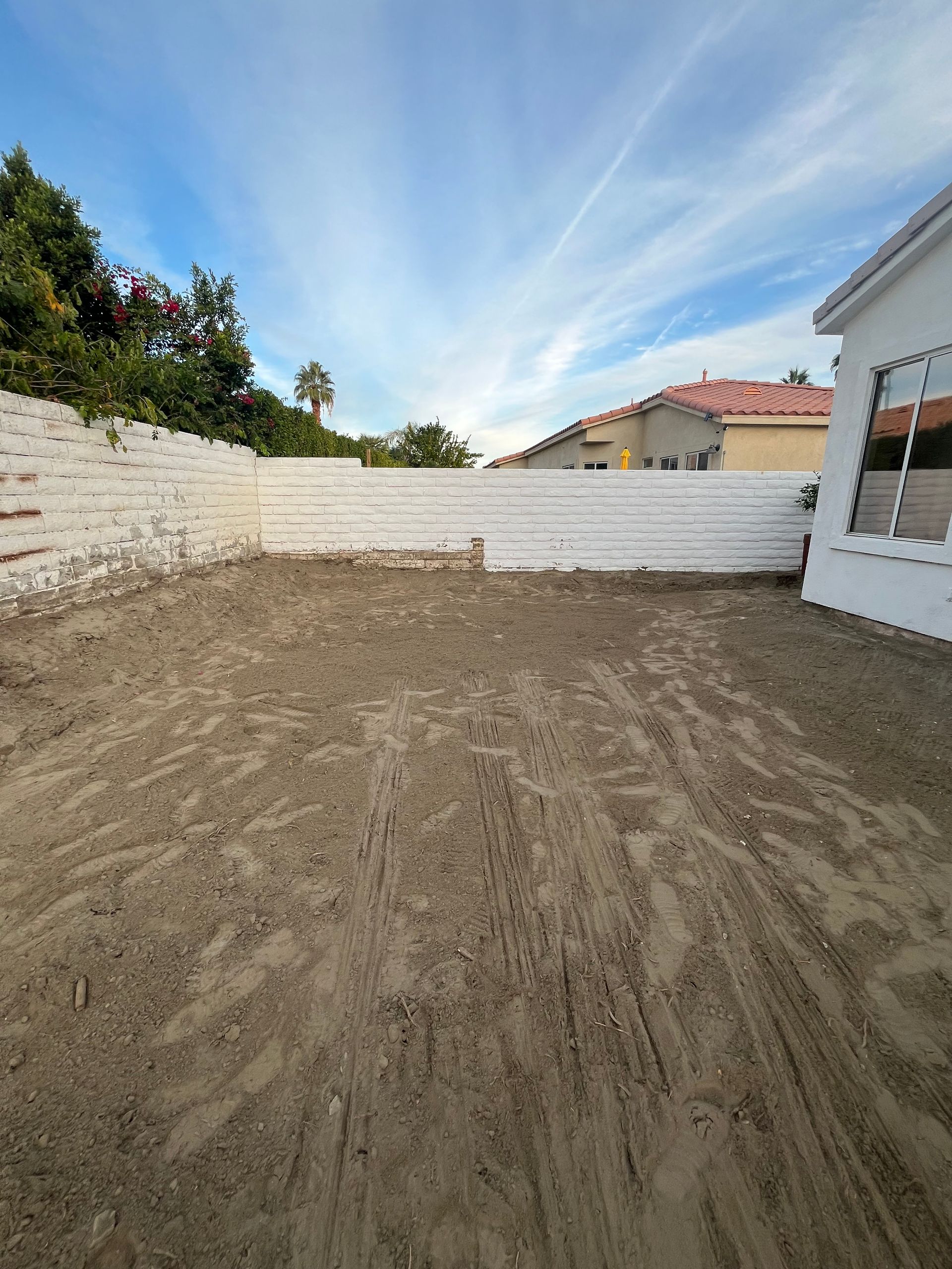 A backyard with a dirt ground and white brick walls under a cloudy blue sky.