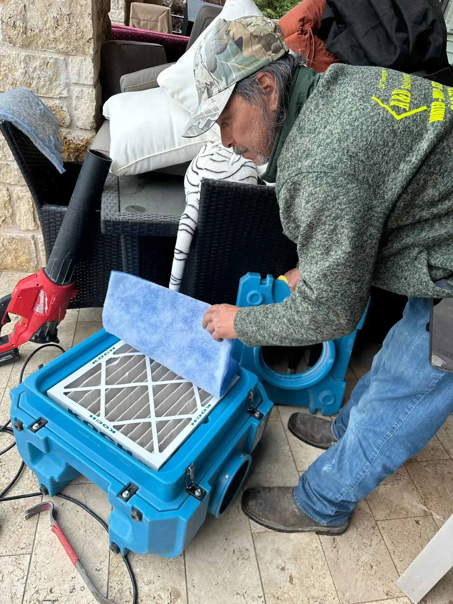 Man replacing an air filter on a blue machine outdoors; other person in the background with a leaf blower.