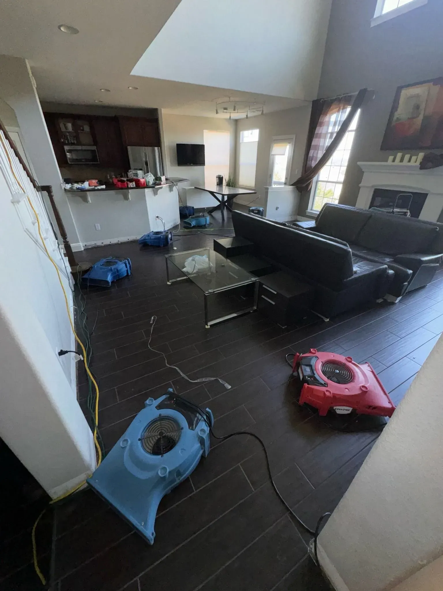 Floor drying equipment in a living room, hardwood floor, black sofa, fireplace, and open kitchen.