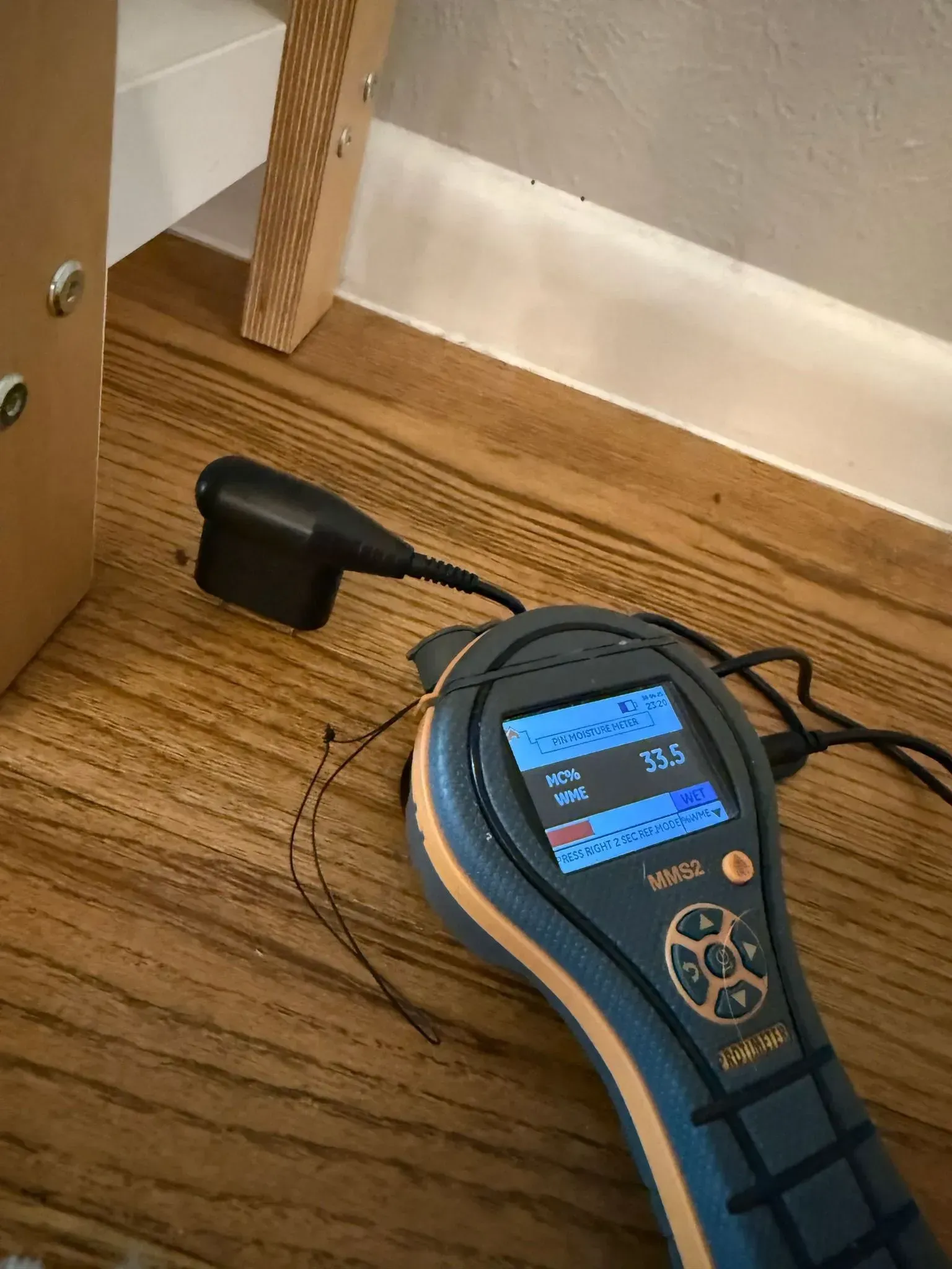 A moisture meter on a hardwood floor, next to a white wall and wooden structure.