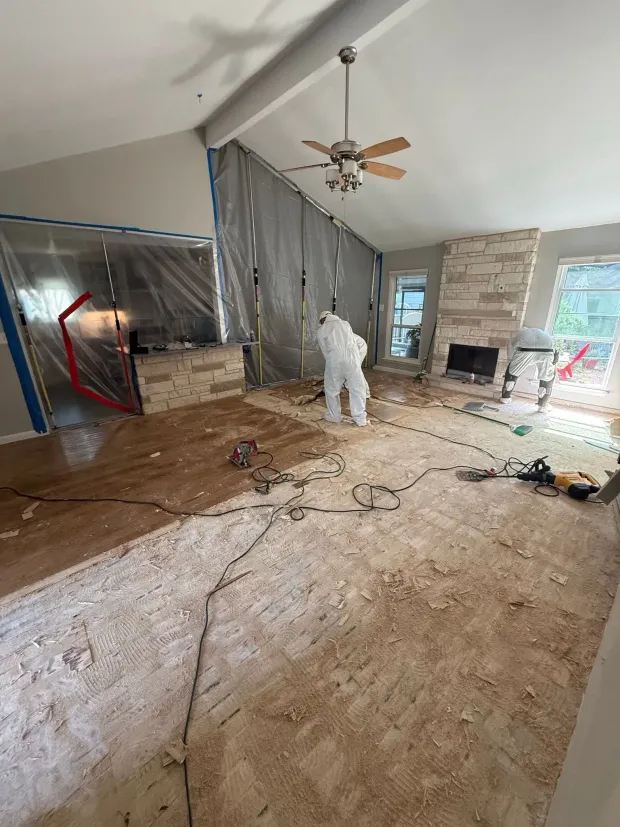 Person in hazmat suit amid construction debris inside a house, tarped walls, stone fireplace.