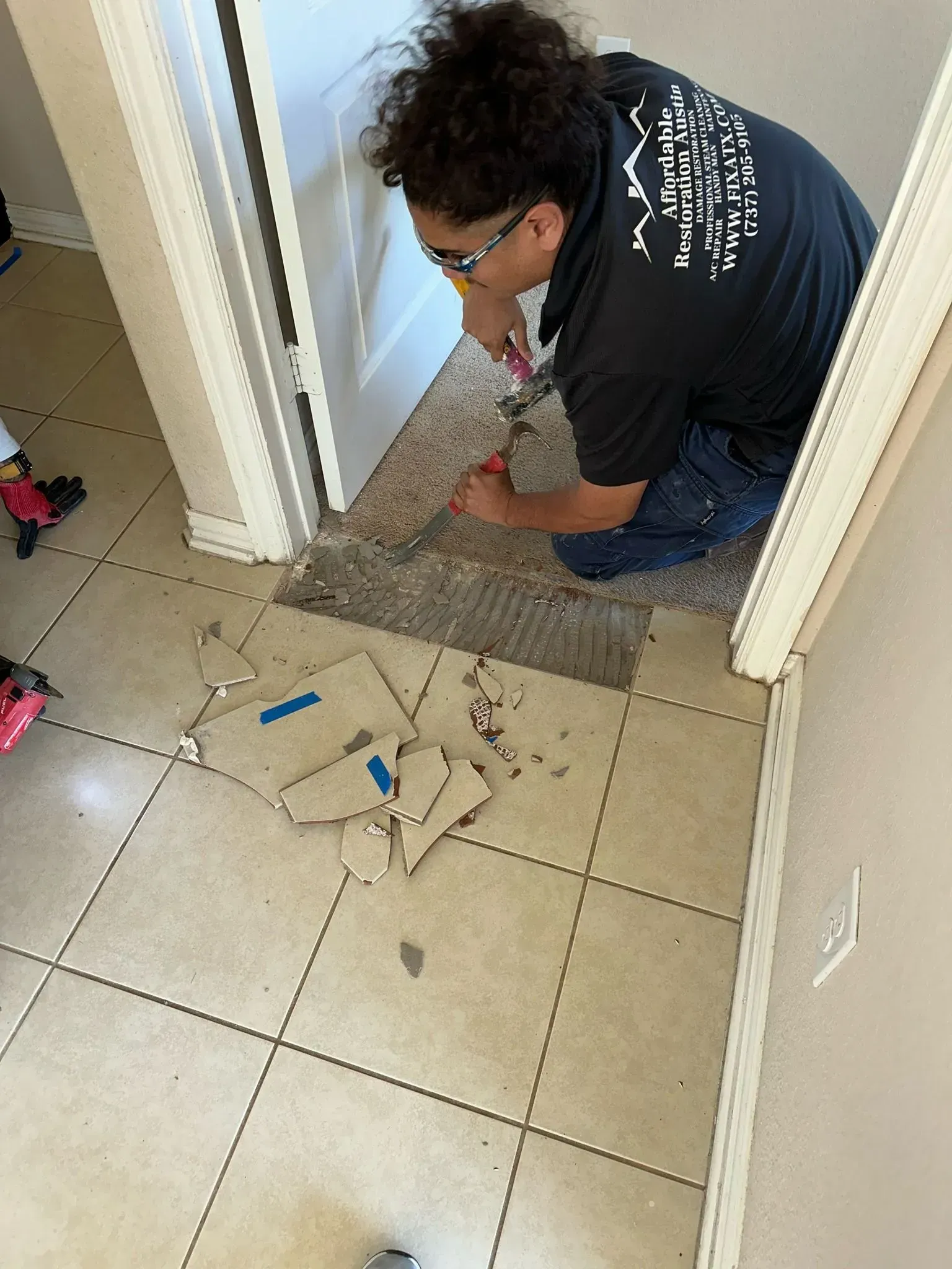 Person kneeling, removing floor tiles in doorway; debris scattered.