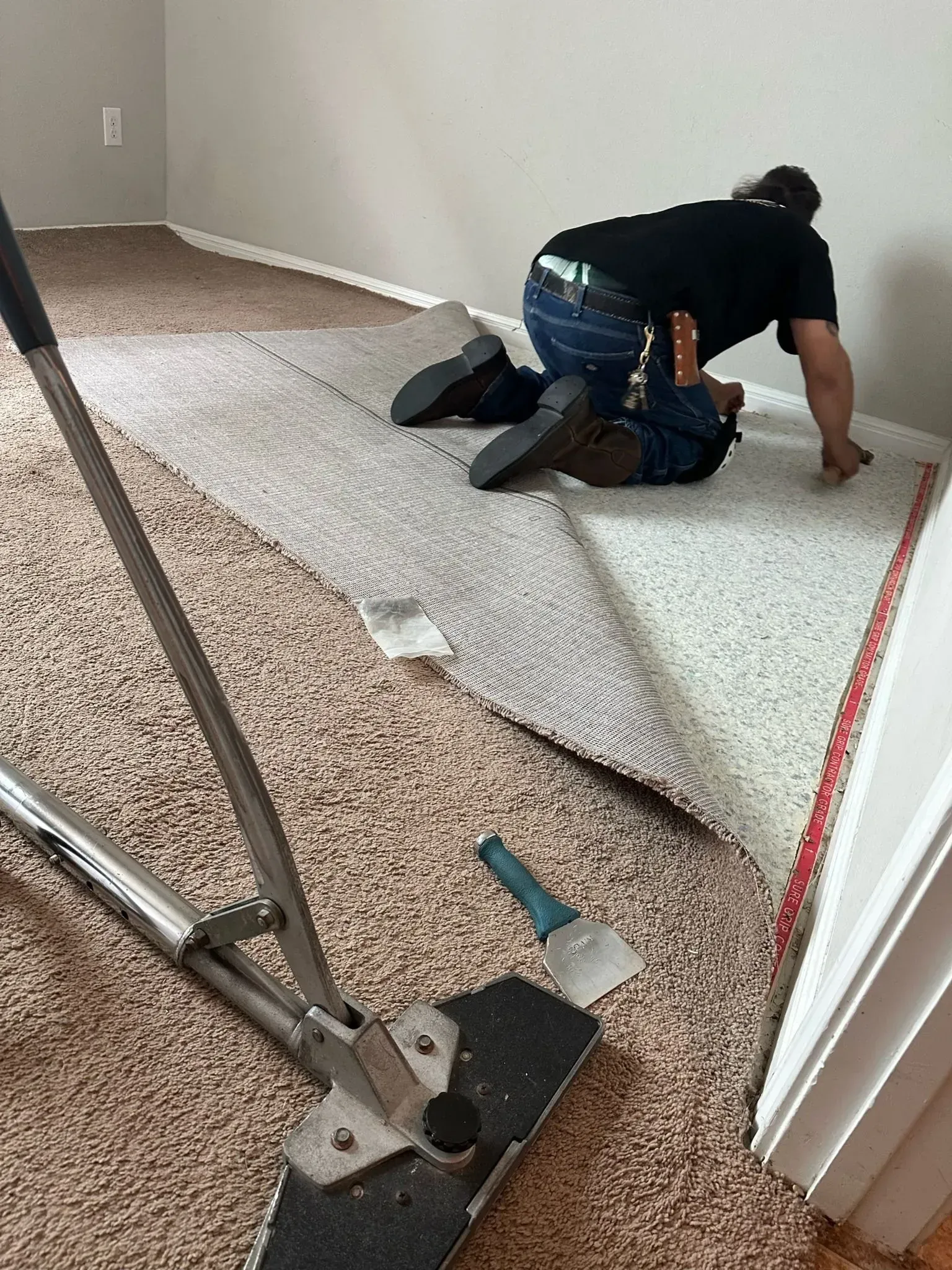 Person installing carpet. One is kneeling. Large cutting tool in foreground. Beige and white carpet.