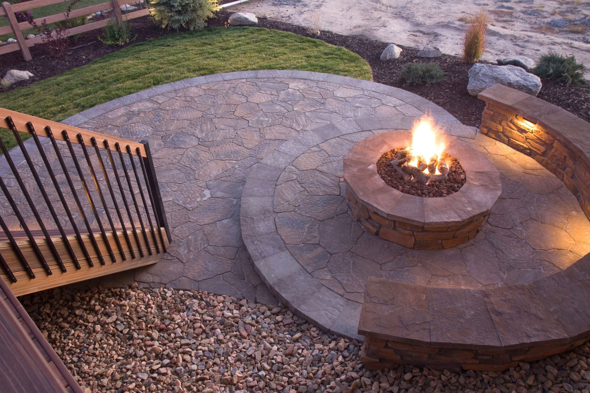 A stone fire pit on a circular patio with an integrated curved seating wall, stairs, and landscaping at twilight.