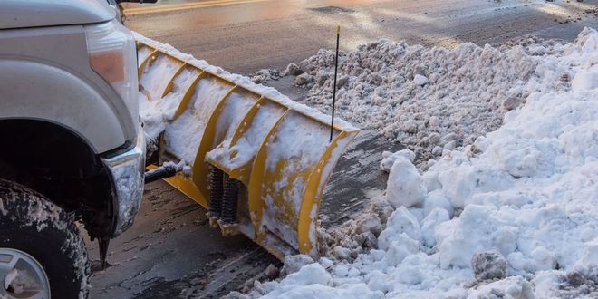 A yellow snowplow blade attached to a vehicle pushing a large pile of snow off a paved road.