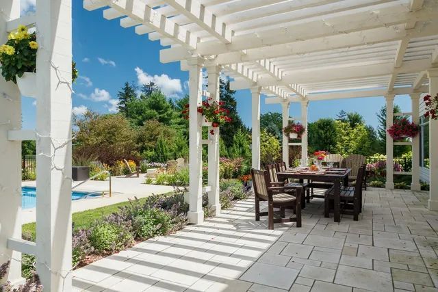 A white wooden pergola covers a stone patio with a dining set, overlooking a swimming pool and landscaped backyard.