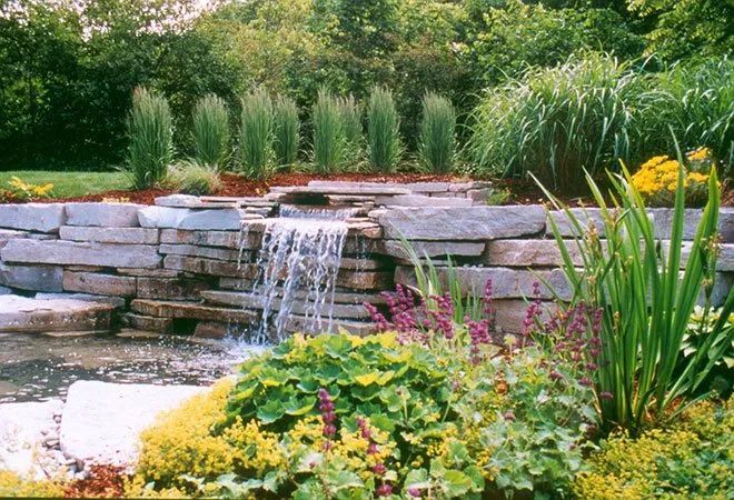 A stone waterfall feature flows into a pond, surrounded by lush green grasses, flowering plants, and trees.