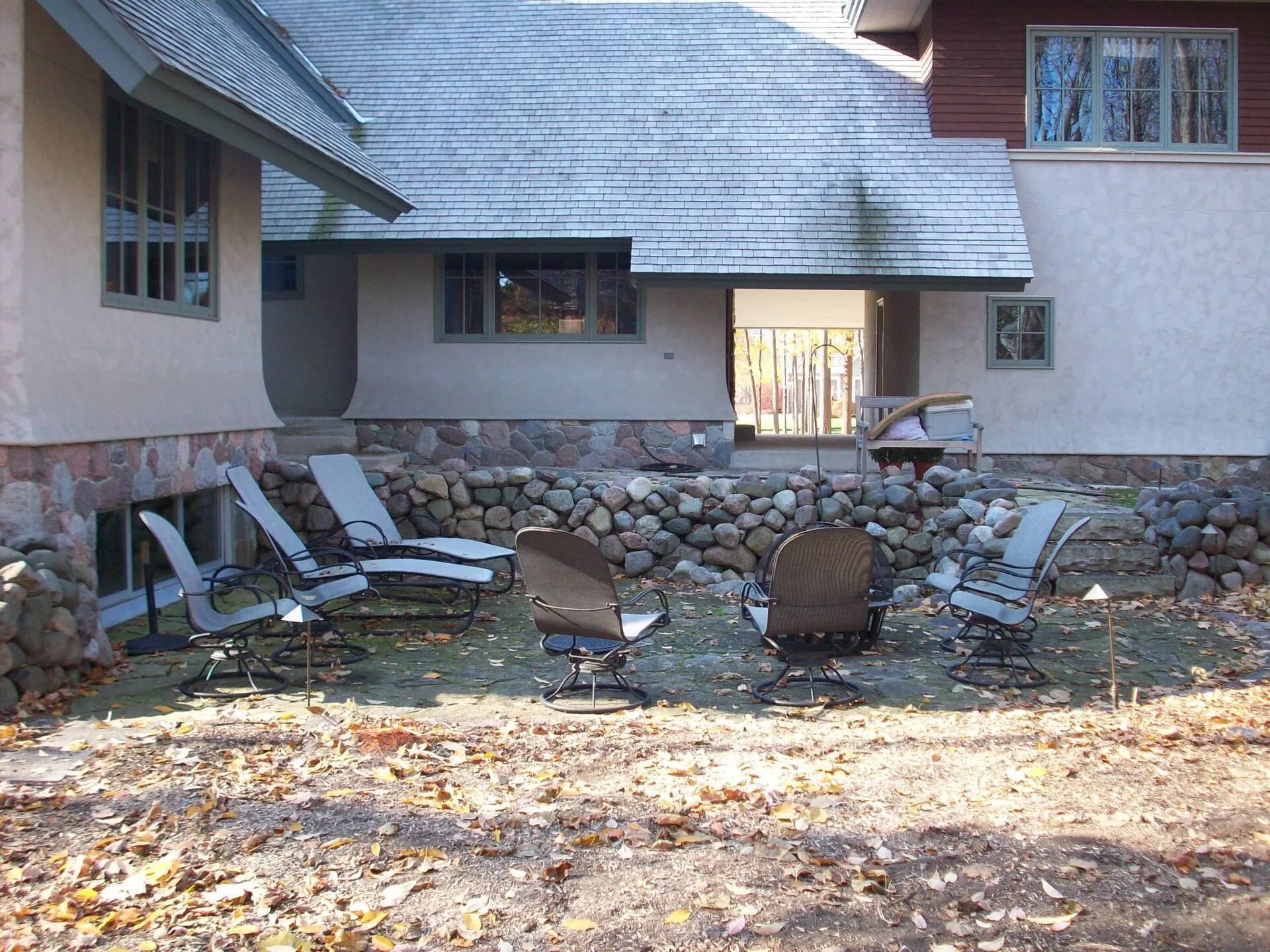 Outdoor patio with stone wall and several chairs in front of a house with a large overhanging roof.