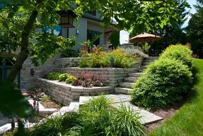 Stone steps lead up a terraced garden featuring tiered stone retaining walls and lush green plantings under tree shade.