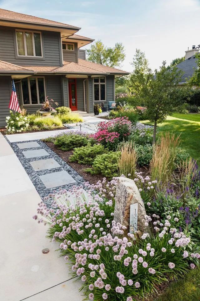 A grey house with a red front door, featuring a path of stone pavers surrounded by a garden of flowers and greenery.