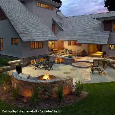 A stone fire pit on a backyard patio at dusk, featuring lounge seating, landscape lighting, and a multi-level modern home.