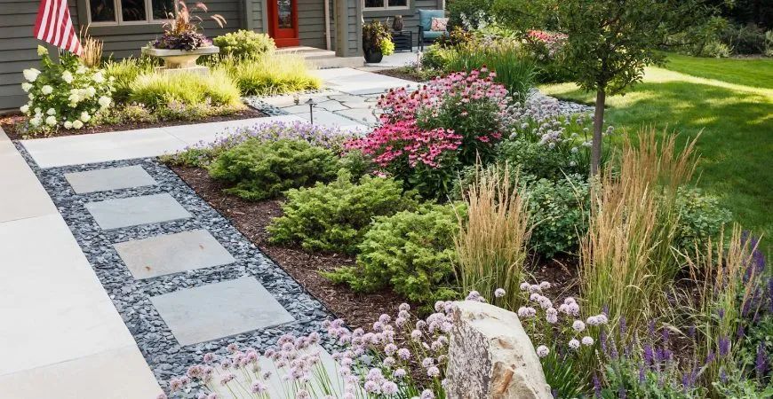 A stone walkway with square pavers and dark gravel leads to a house entrance, surrounded by colorful perennial gardens.