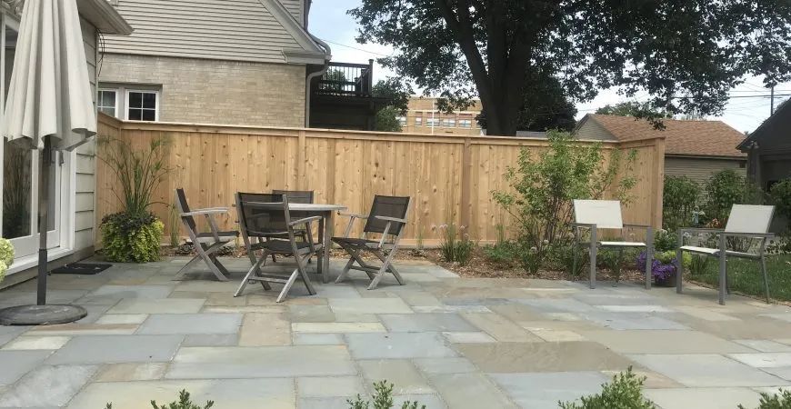 A patio with stone pavers, a dining set with four chairs, and two separate chairs near a wooden privacy fence.