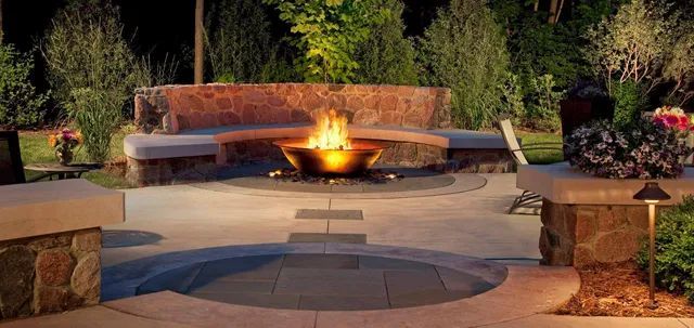 A curved stone fire pit with a glowing fire sits in a patio garden with integrated seating under night lighting.